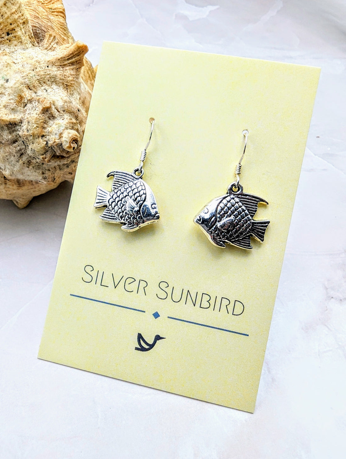Displayed on a yellow card, the "Angelic Angelfish Earrings" by Silver Sunbird epitomize ocean-themed elegance. Set against a light-coloured surface with a seashell in the background, these sterling silver earrings are perfect for any ocean lover.