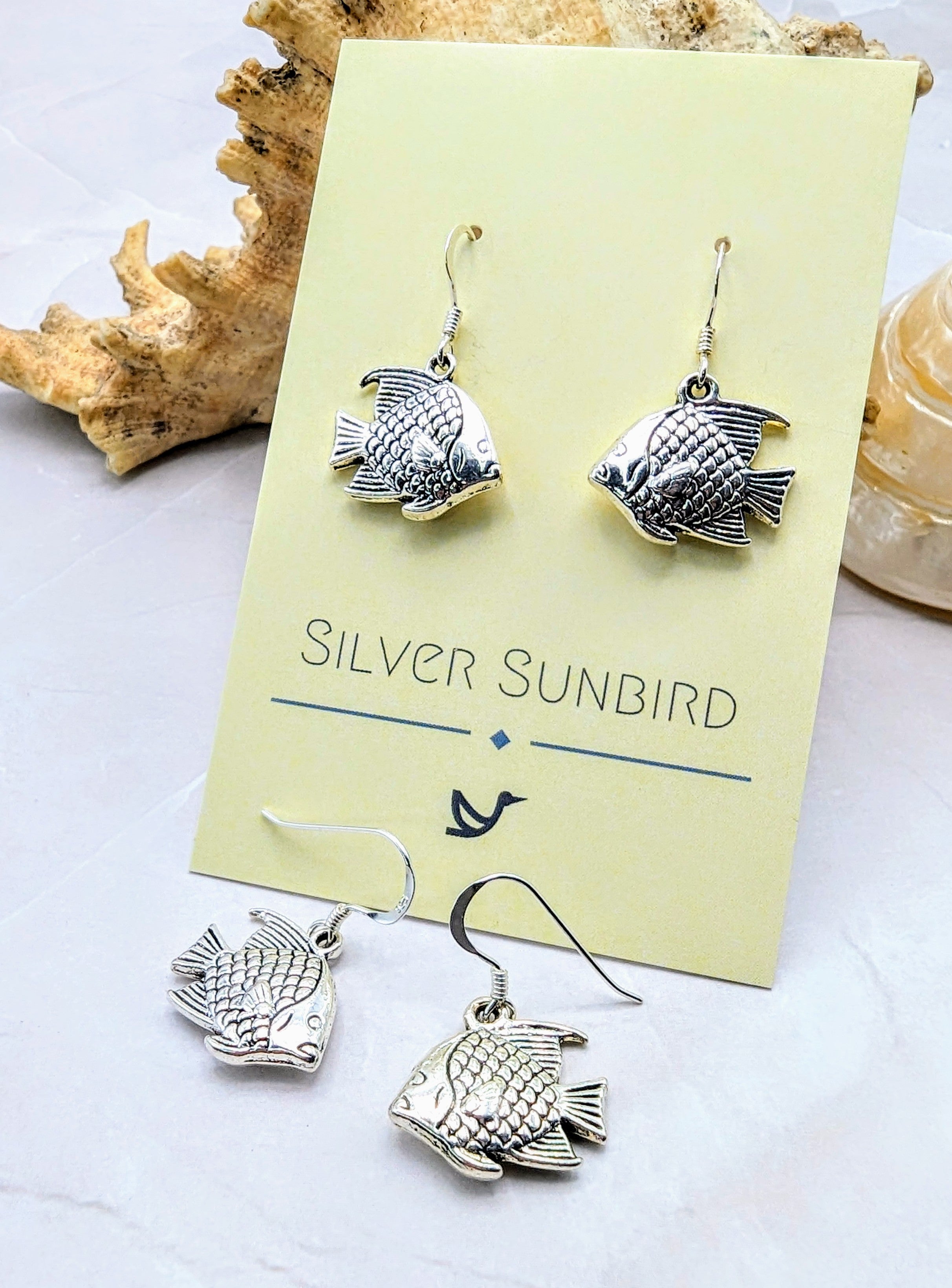A pair of Angelic Angelfish Earrings, crafted from Sterling silver and perfect for any ocean lover, are displayed on a yellow card with the brand name "Silver Sunbird." Behind the card is a piece of coral, while another identical pair of these exquisite earrings is laid out in front.