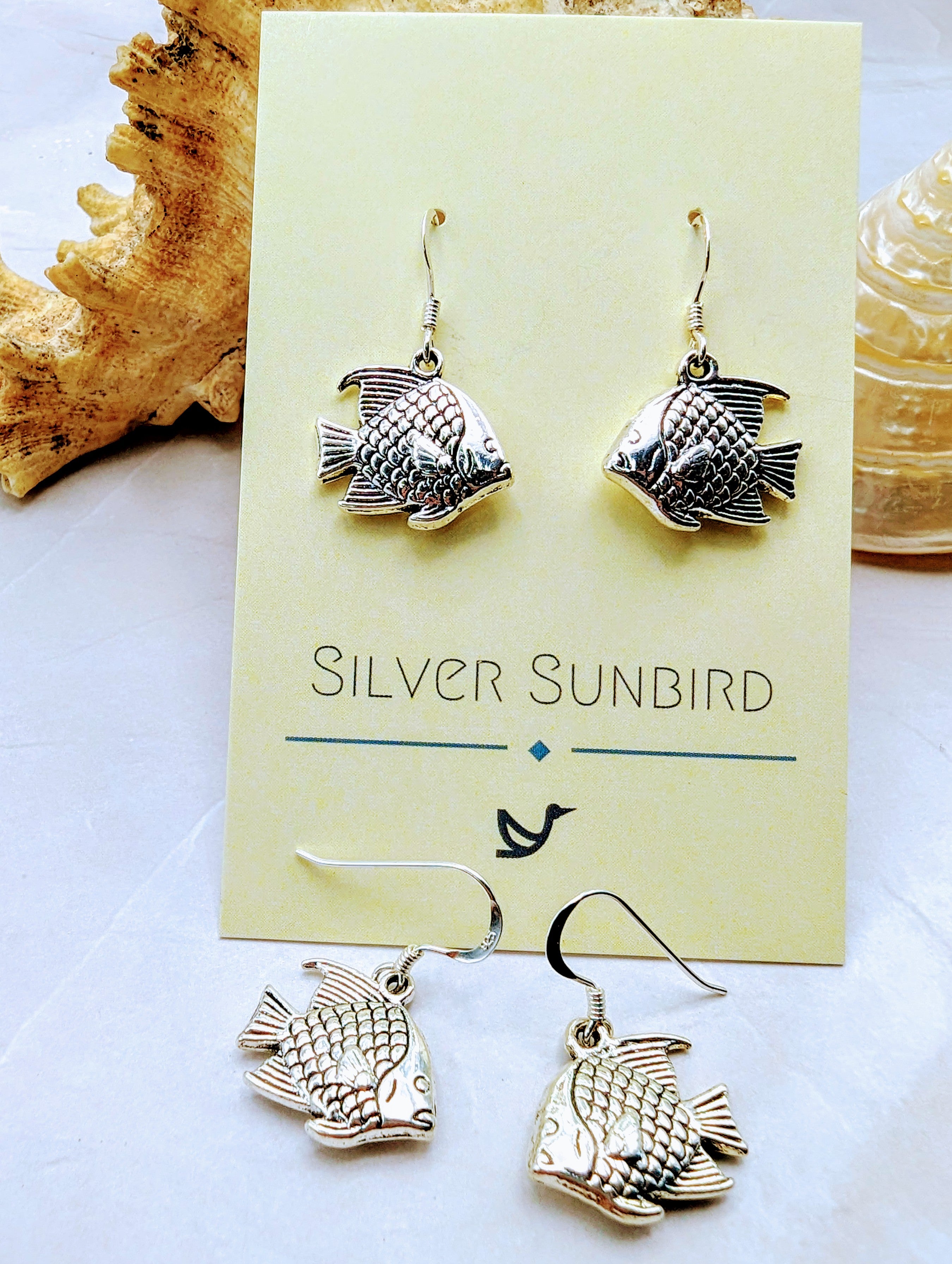 A display of the Silver Sunbird Angelic Angelfish Earrings is showcased. Two sterling silver earrings are attached to a branded card, while another pair is laid out on the surface below. There is a textured decorative item in the background, slightly out of focus, perfect for any ocean lover.