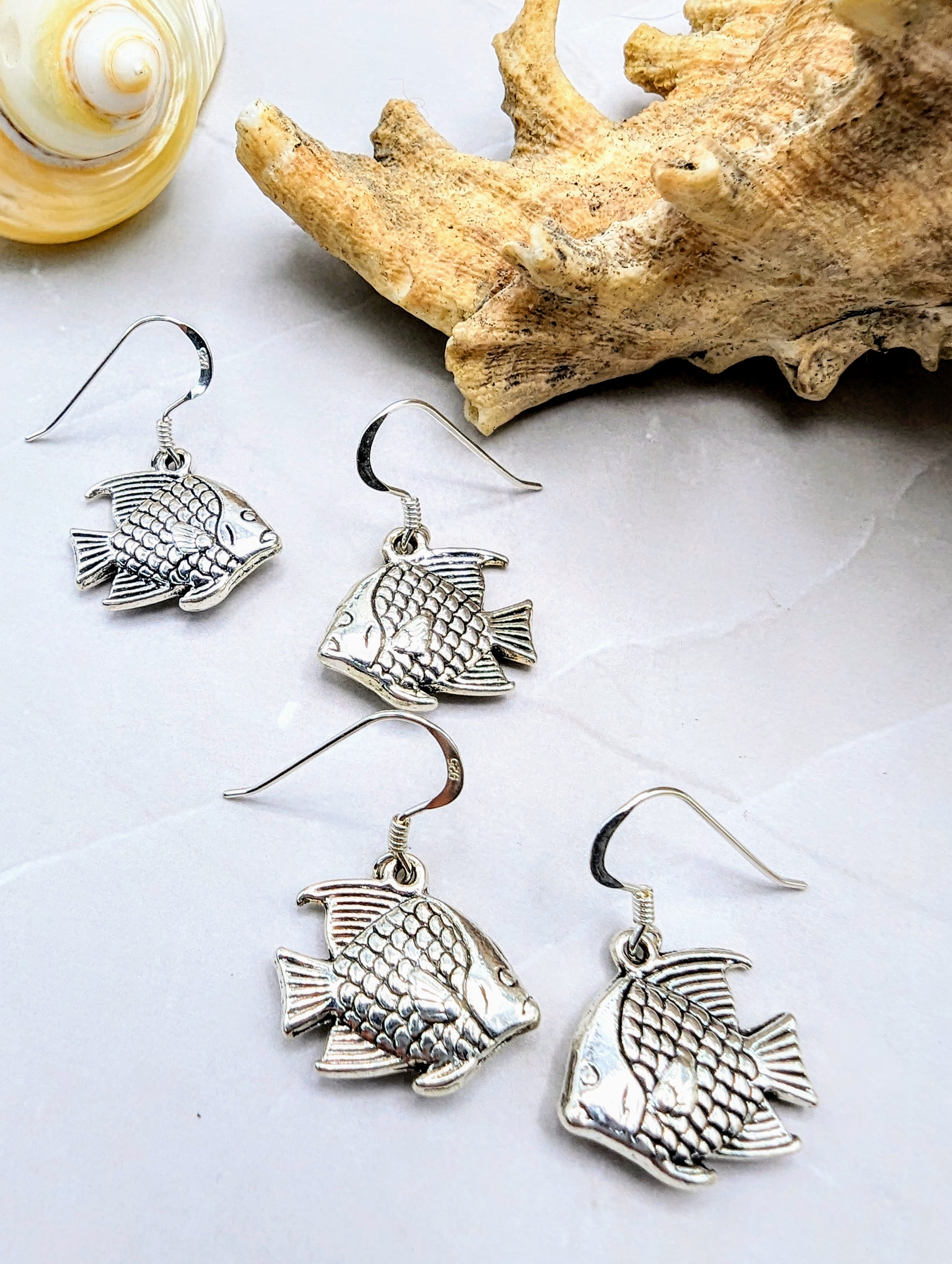 A set of four Angelic Angelfish Earrings by Silver Sunbird displayed on a grey surface, accompanied by a seashell and a piece of coral for decoration. The sterling silver earrings feature detailed scales and fins, each with a hook for wearing. Perfect for any ocean lover, the setting suggests an oceanic or beach-themed style.