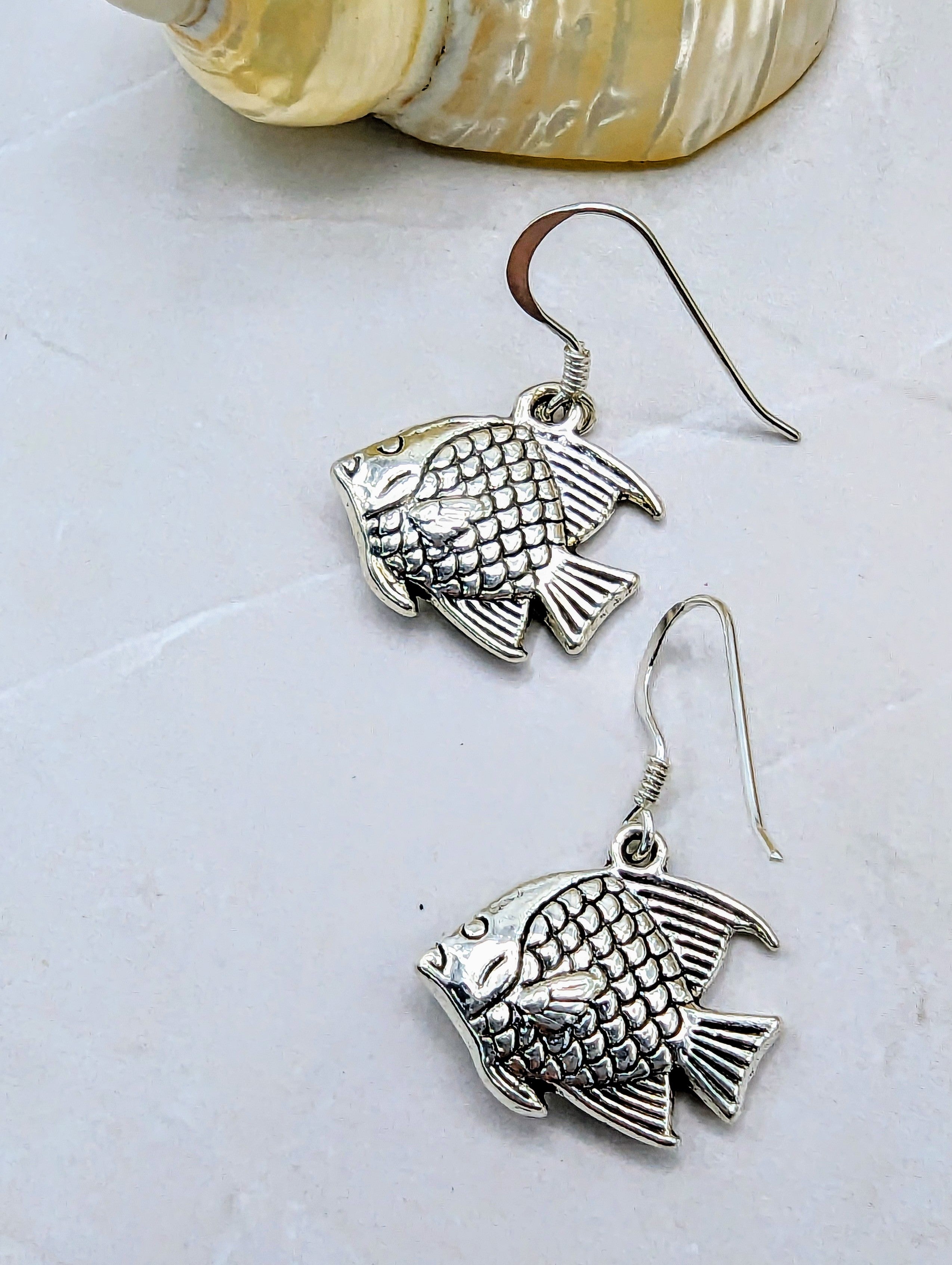 The Angelic Angelfish Earrings by Silver Sunbird, crafted from sterling silver and featuring intricately detailed scales and fins, are displayed on a light, textured surface. Perfect for any ocean lover, these angelfish earrings have hook-style backs and include part of a shell visible in the upper part of the image.