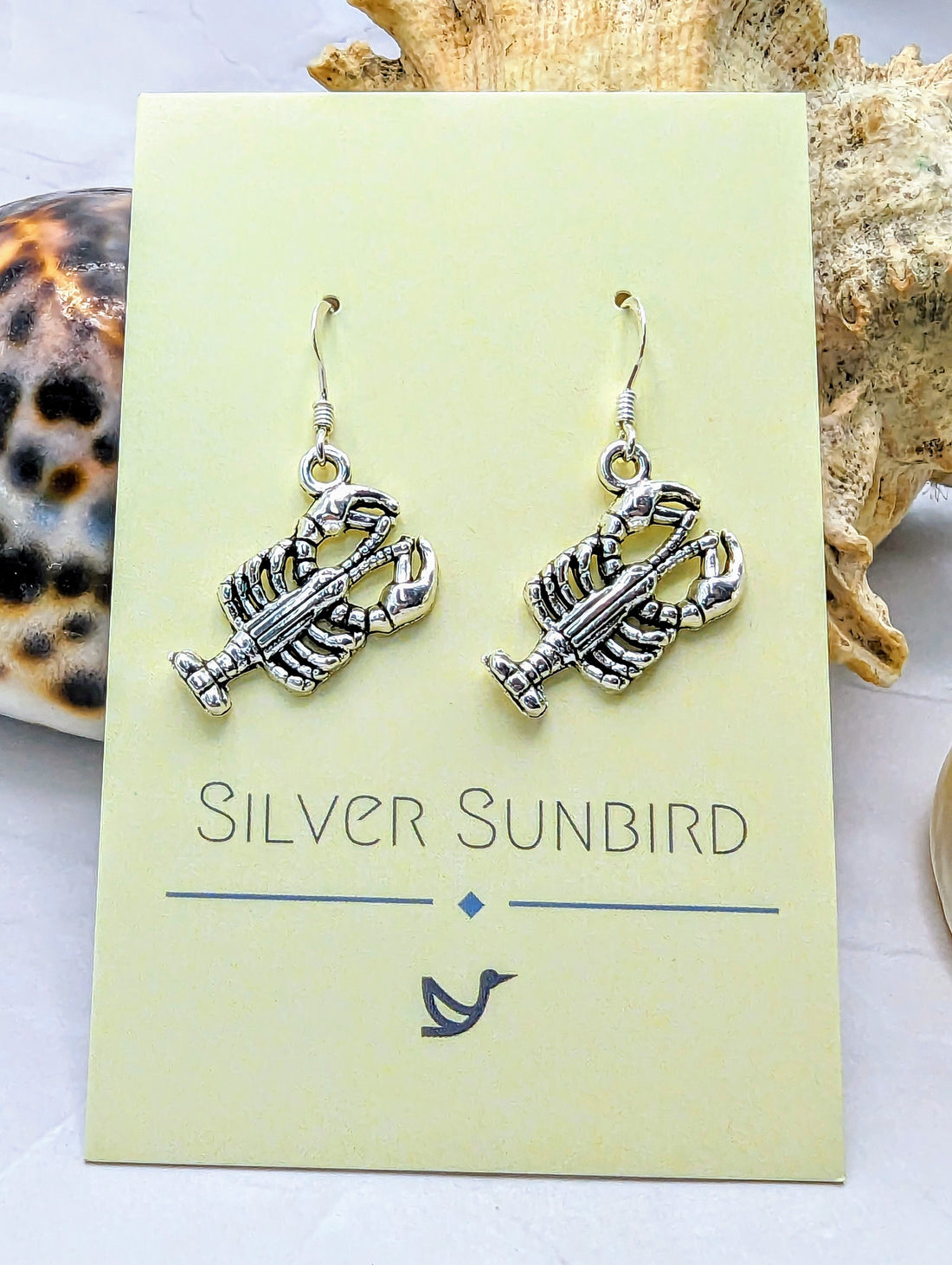 A pair of hypoallergenic, silver lobster-shaped Lively Lobster Earrings by Silver Sunbird is displayed on a card. The seaside-themed presentation, with seashell and conch decor, captures an ocean spirit.