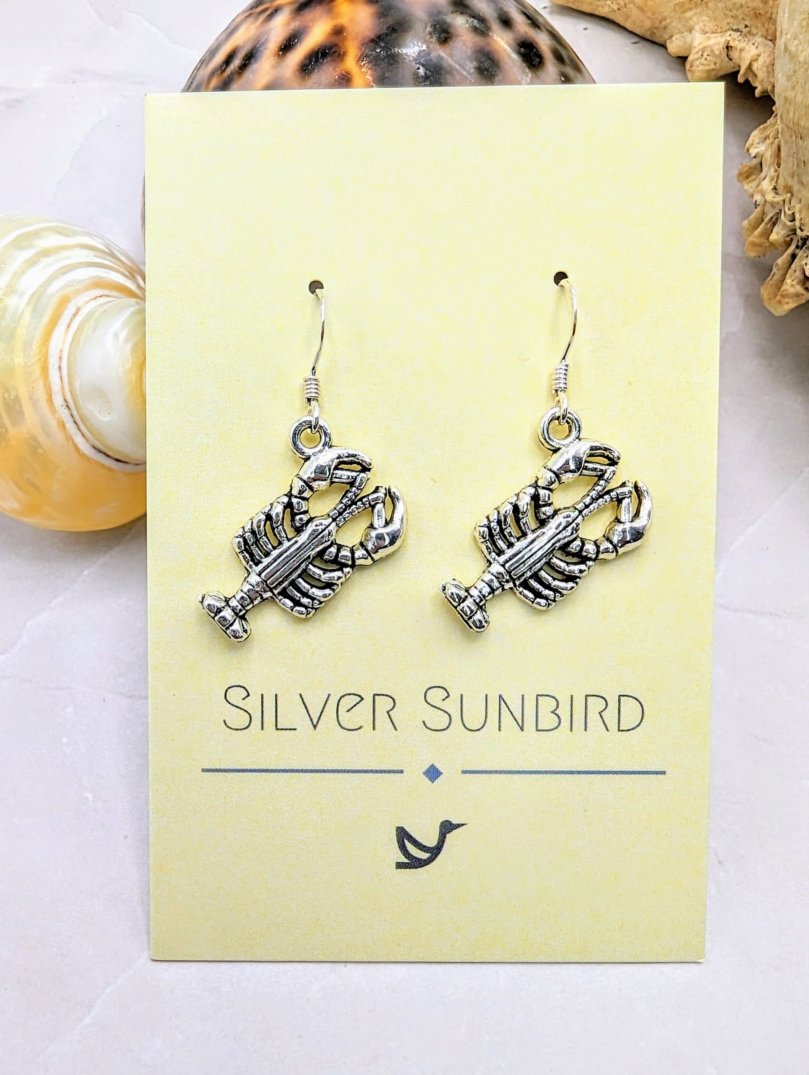 The Lively Lobster Earrings by Silver Sunbird channel ocean vibes on a card labeled "Silver Sunbird," elegantly placed on a light stone surface with decorative shells. These earrings are hypoallergenic and feature a lobster shape.