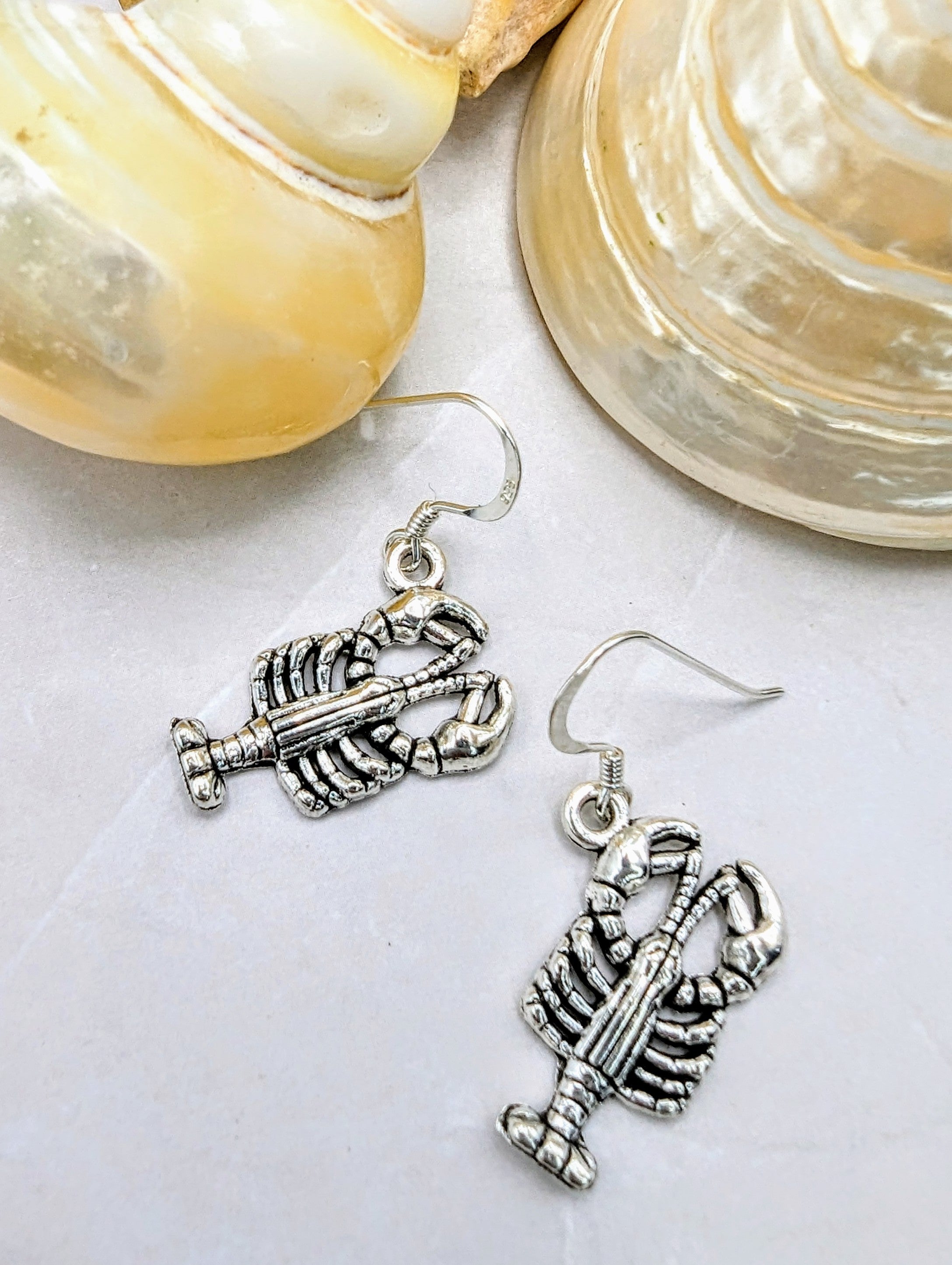 Two hypoallergenic Silver Sunbird Lively Lobster Earrings with intricate detailing lie on a light surface beside two large, cream-colored seashells. With hooks for wearing, they evoke an ocean spirit and are perfect for those with sensitive skin.