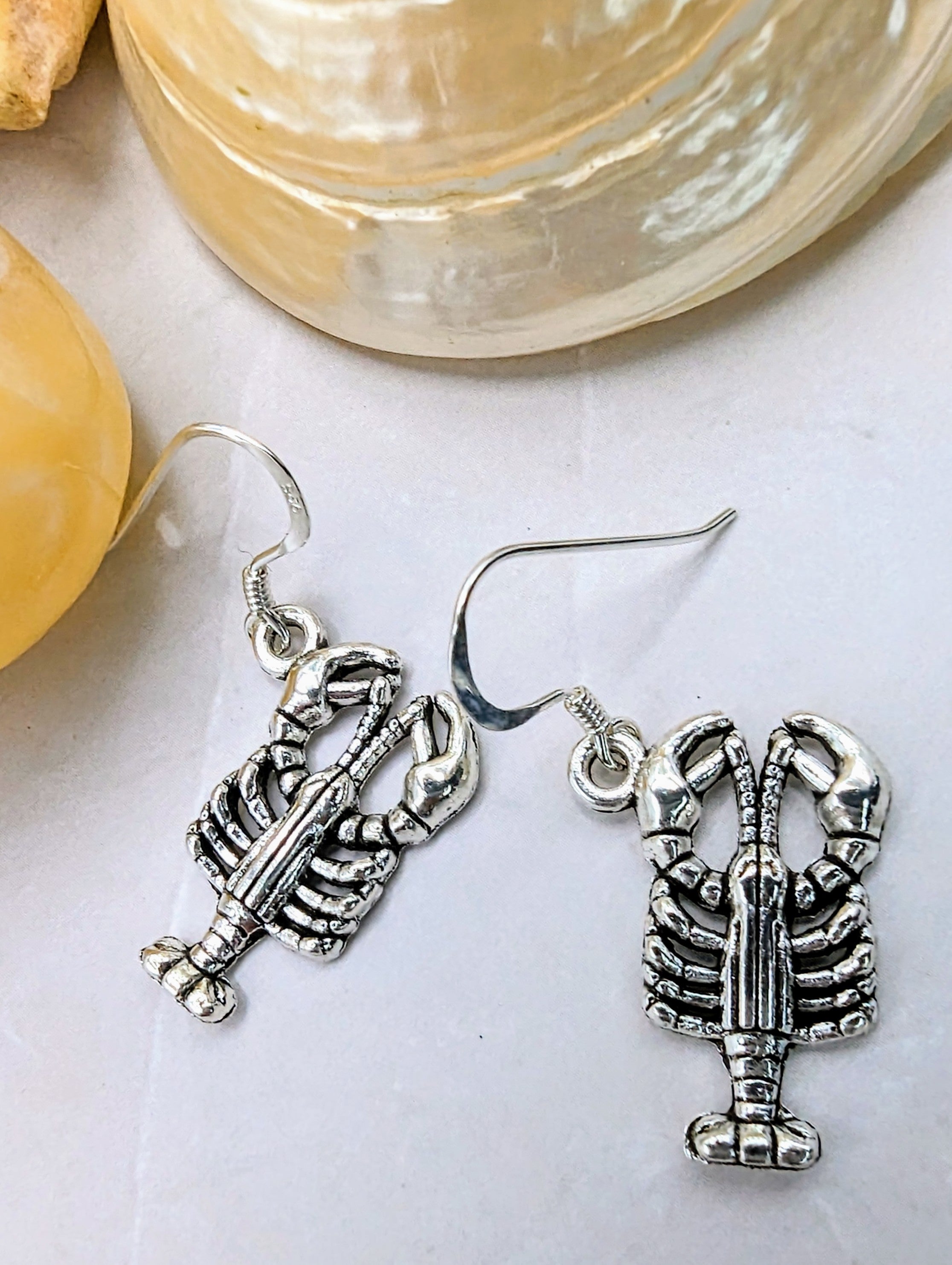 The Silver Sunbird Lively Lobster Earrings, hypoallergenic and precisely detailed to capture the ocean spirit, are displayed on a white surface surrounded by seashells.