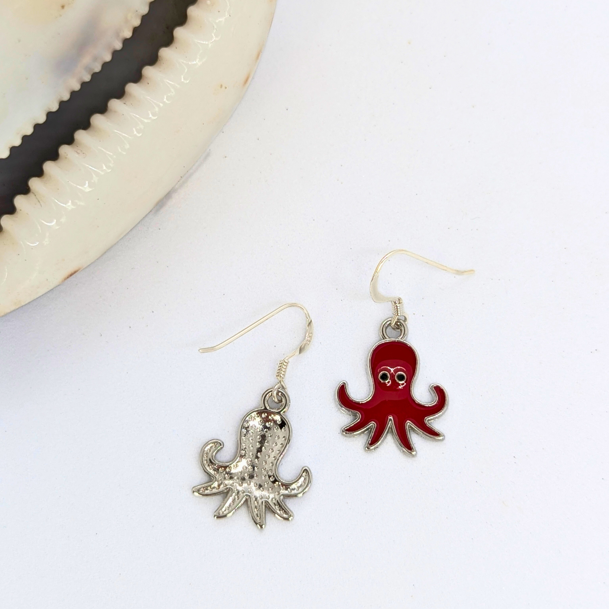 Two ocean-inspired earrings from Silver Sunbird—one a silver octopus, the other a Red Octopus Earring with enamel and eyes—are displayed beside a white seashell on a white background.