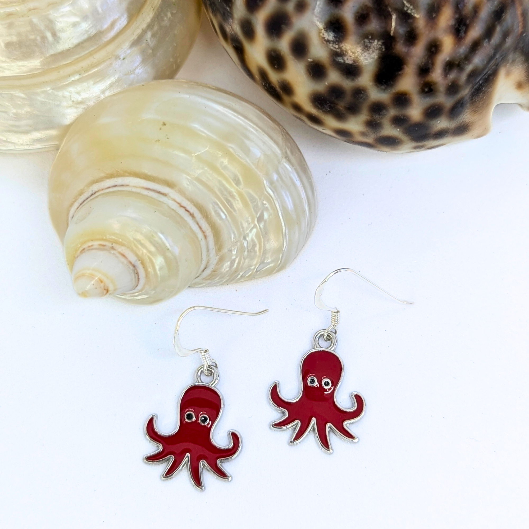 Two ocean-inspired Red Octopus Earrings by Silver Sunbird, featuring googly eyes and red enamel on sterling silver, rest on a white surface next to a spiral cream seashell and a spotted dark seashell.