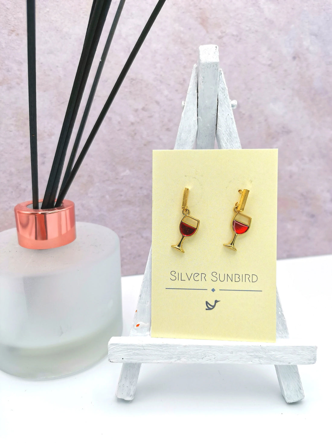 Red Wine Drop Studs by Silver Sunbird—handmade, hypoallergenic gold earrings shaped like wine glasses with red liquid, displayed on a branded card set on a white easel beside a diffuser with black reeds and rose gold rim.