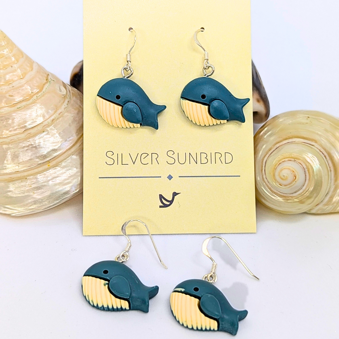 A pair of Colourful Whale Earrings by Silver Sunbird is displayed on a branded card with two seashells beside them, while another identical sterling silver whale earring pair lies in front.