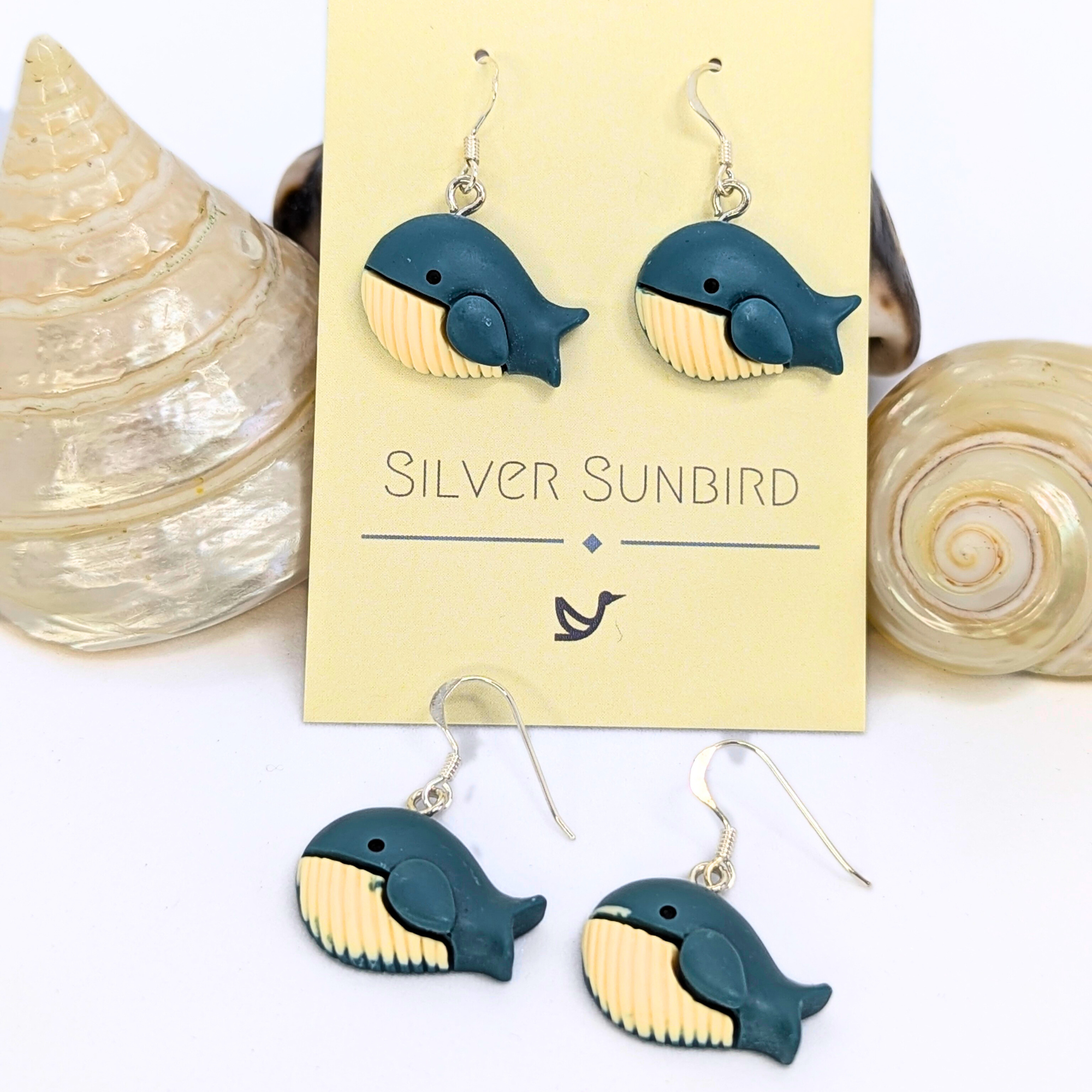Colourful Whale Earrings by Silver Sunbird, shaped like blue whales with beige bellies, displayed on a branded card and accented by spiral seashells—perfect for adding coastal charm to your style.
