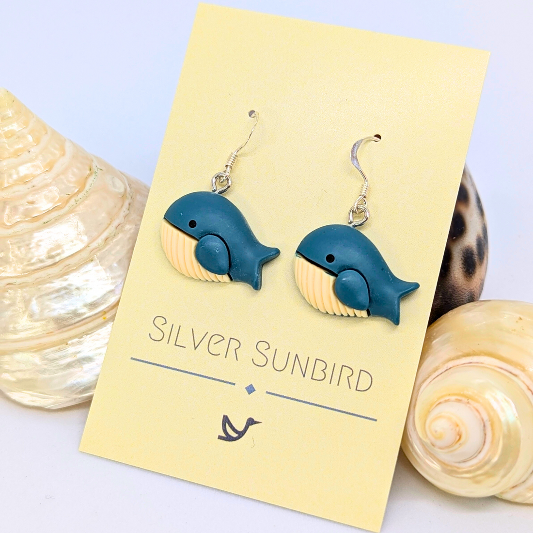 Colourful Whale Earrings by Silver Sunbird—blue and cream earrings displayed on a branded card, surrounded by seashells. The perfect nautical accessory for ocean lovers.