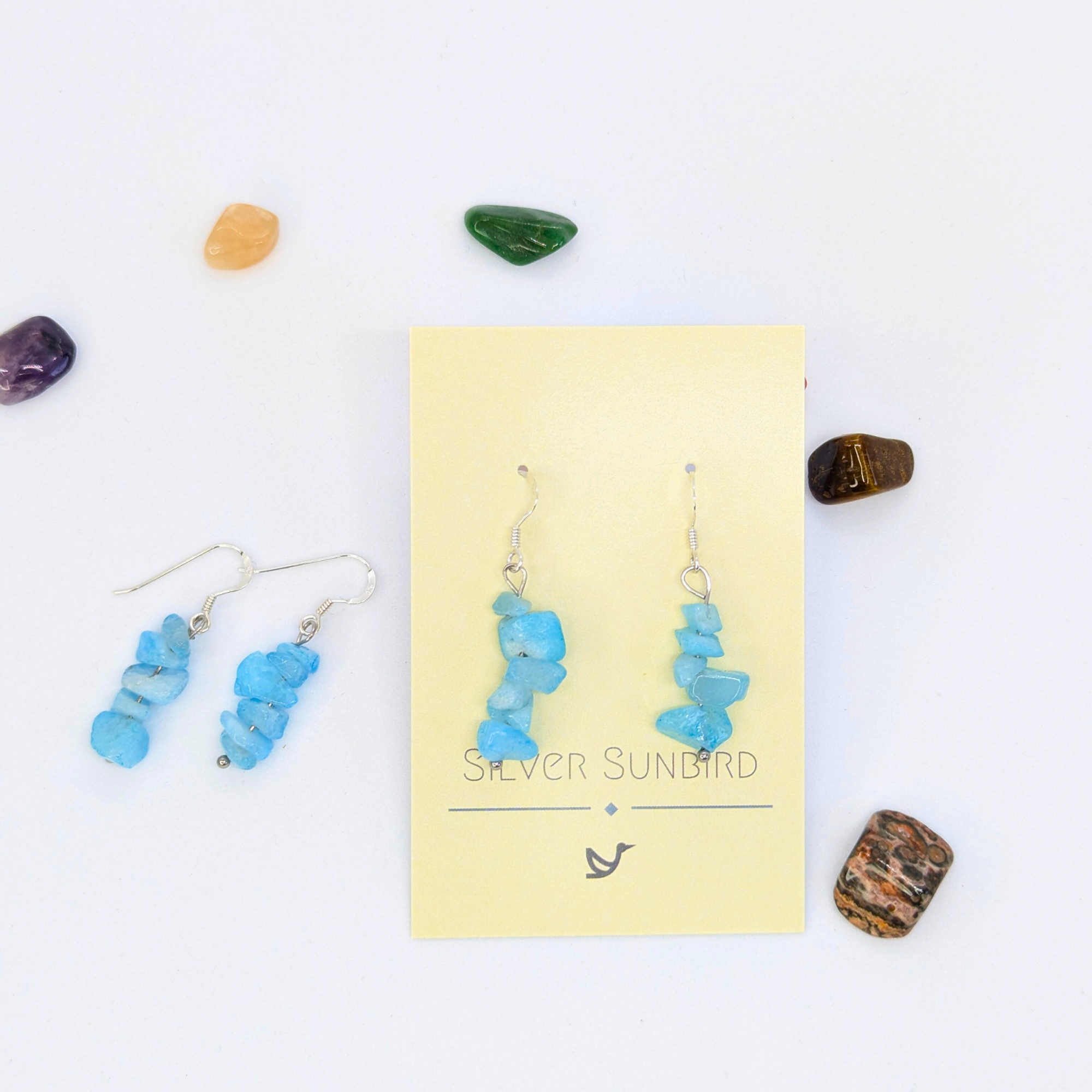 Aquamarine Gemstone Earrings by Silver Sunbird displayed on a yellow branded card, surrounded by colorful stones. A matching pair of sterling silver hypoallergenic earrings rests to the left on a white background.