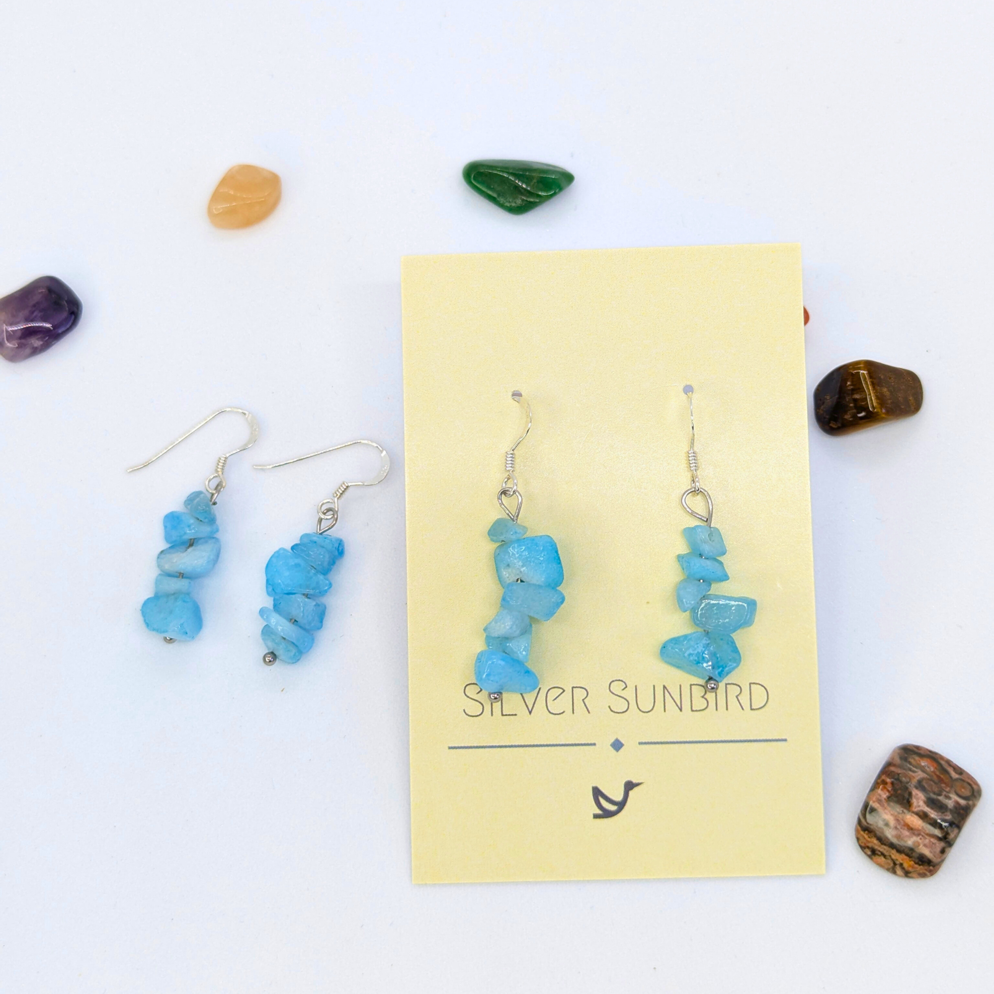 Aquamarine Gemstone Earrings by Silver Sunbird displayed on a yellow brand card, surrounded by colorful polished stones on a white background.