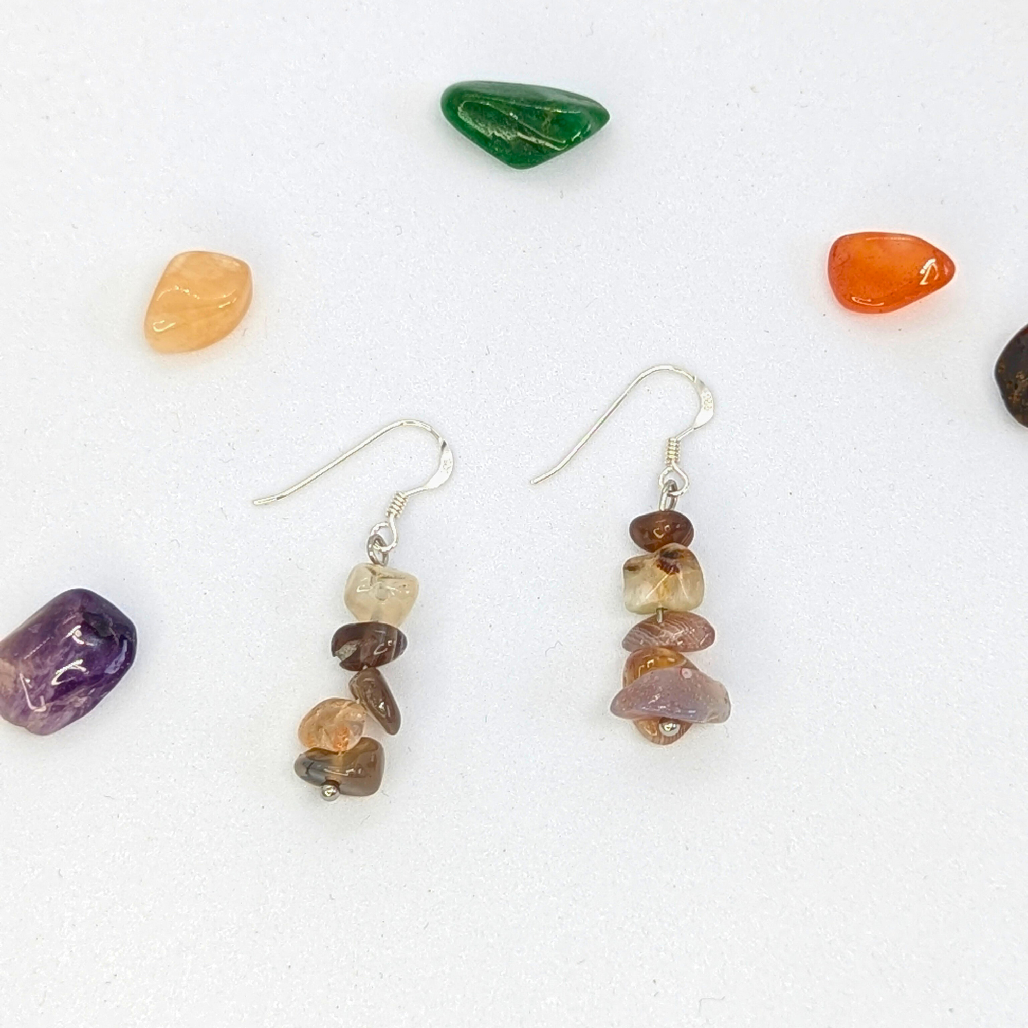 The Silver Sunbird Botswana Agate Gemstone Earrings feature stacked brown and beige gemstone chips, displayed on a white surface and surrounded by colorful polished stones.