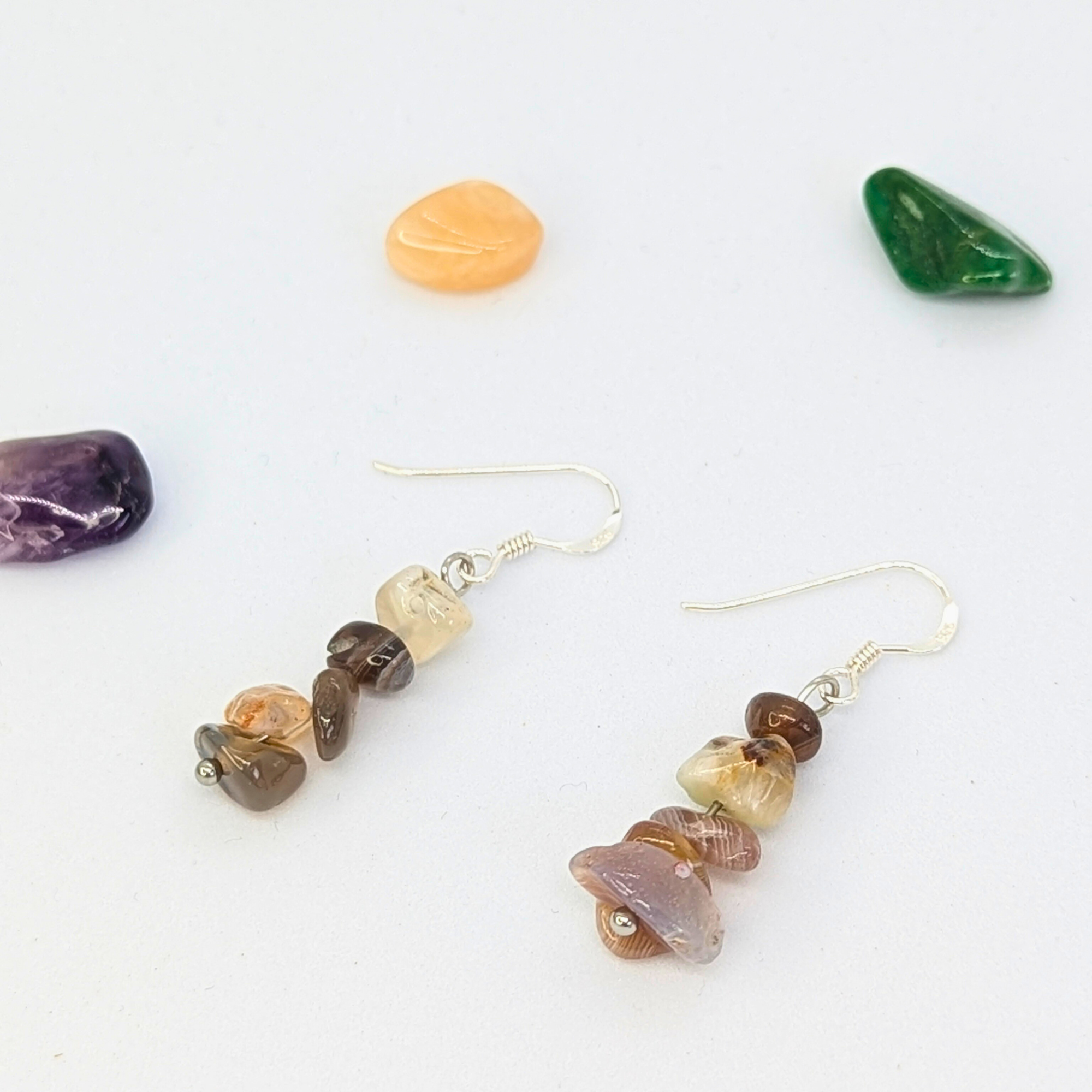 Silver Sunbird’s Botswana Agate Gemstone Earrings, featuring stacked multicolored stone beads, rest on a white surface surrounded by polished green, orange, and purple stones.