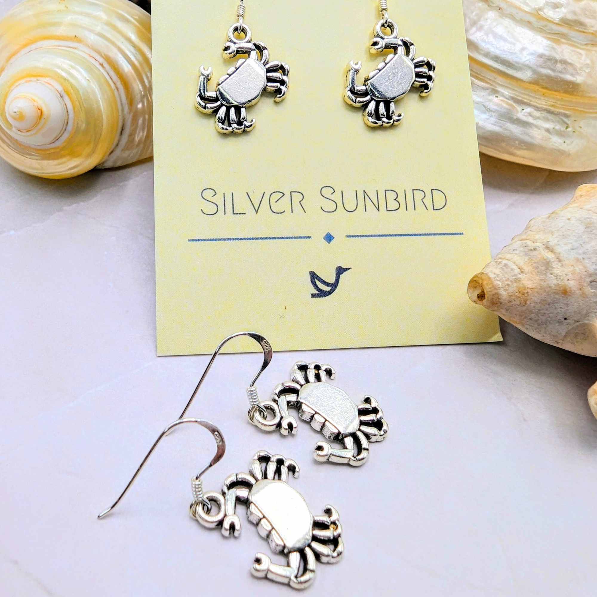 The Coastal Crab Earrings by Silver Sunbird, crafted in sterling silver, are displayed on a branded card amid seashells. These earrings capture seaside charm with their unique crab design.