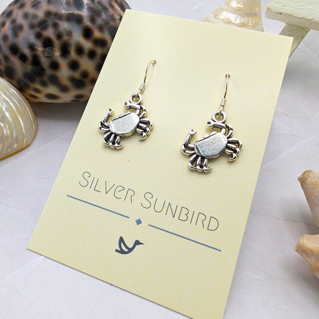 Coastal Crab Earrings by Silver Sunbird are shown on a cream card labeled with the brand name, surrounded by seashells and starfish on a light surface.