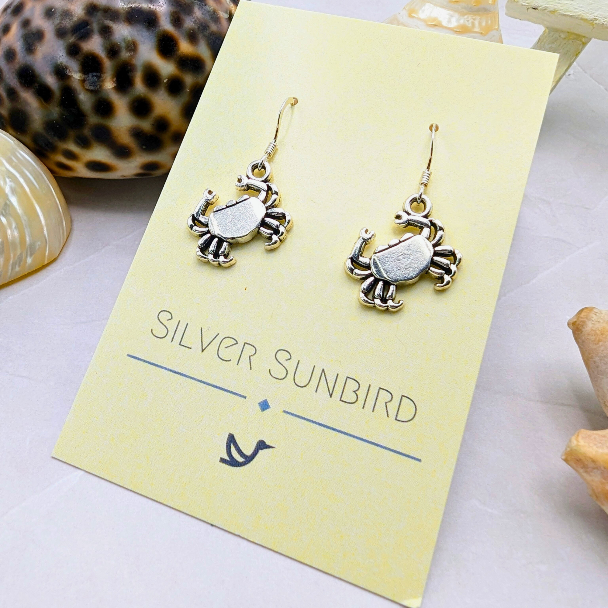 Coastal Crab Earrings by Silver Sunbird—sterling silver dangle earrings shown on a yellow branded card, surrounded by seashells and coral.