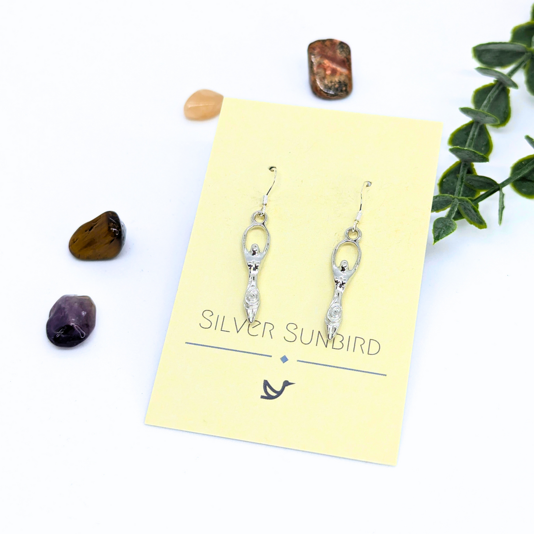 A pair of handmade Fertility Goddess Dangle Earrings by Silver Sunbird, displayed on a yellow branded card and surrounded by assorted stones and greenery against a white background.