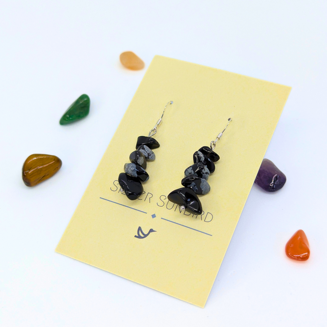 Snowflake Obsidian Earrings by Silver Sunbird dangle elegantly on a yellow card, surrounded by colorful polished stones, highlighting the beauty of handmade crystal jewelry against a crisp white background.
