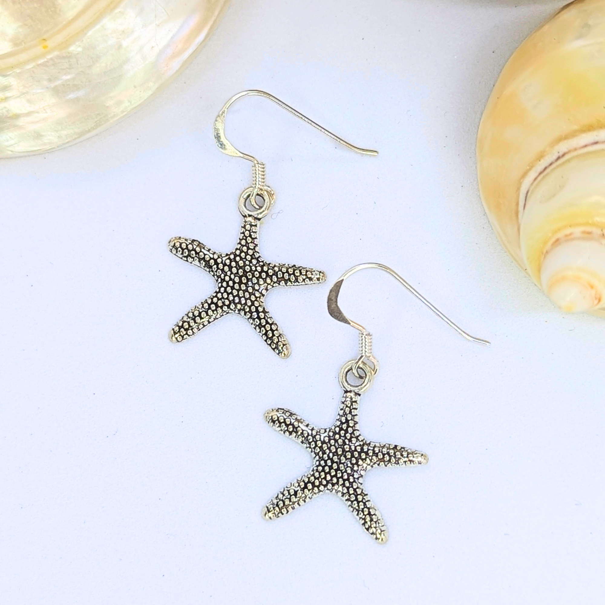 The Silver Sunbird Detailed Silver Starfish Earrings feature textured silver starfish shapes, perfect for boho jewellery lovers, displayed on a white background with two seashells.