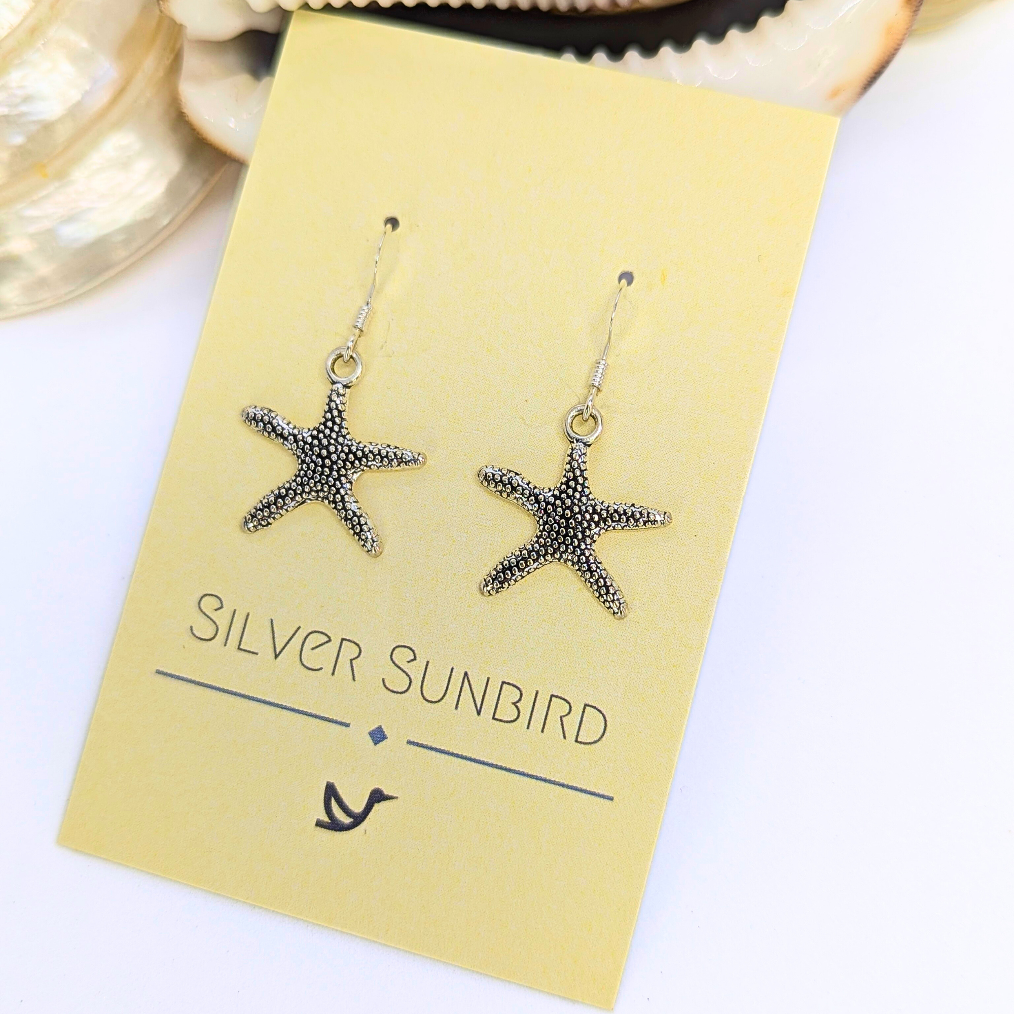 Detailed Silver Starfish Earrings by Silver Sunbird shown on a pale yellow card with a shell nearby—an ideal pick for lovers of unique boho jewellery.
