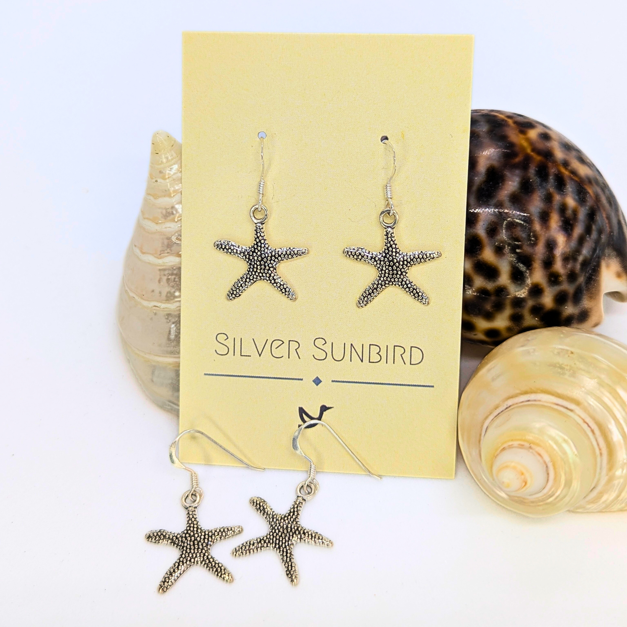 Detailed Silver Starfish Earrings by Silver Sunbird displayed on a branded card, with three seashells on a white surface—perfect boho jewellery for a seaside-inspired look.