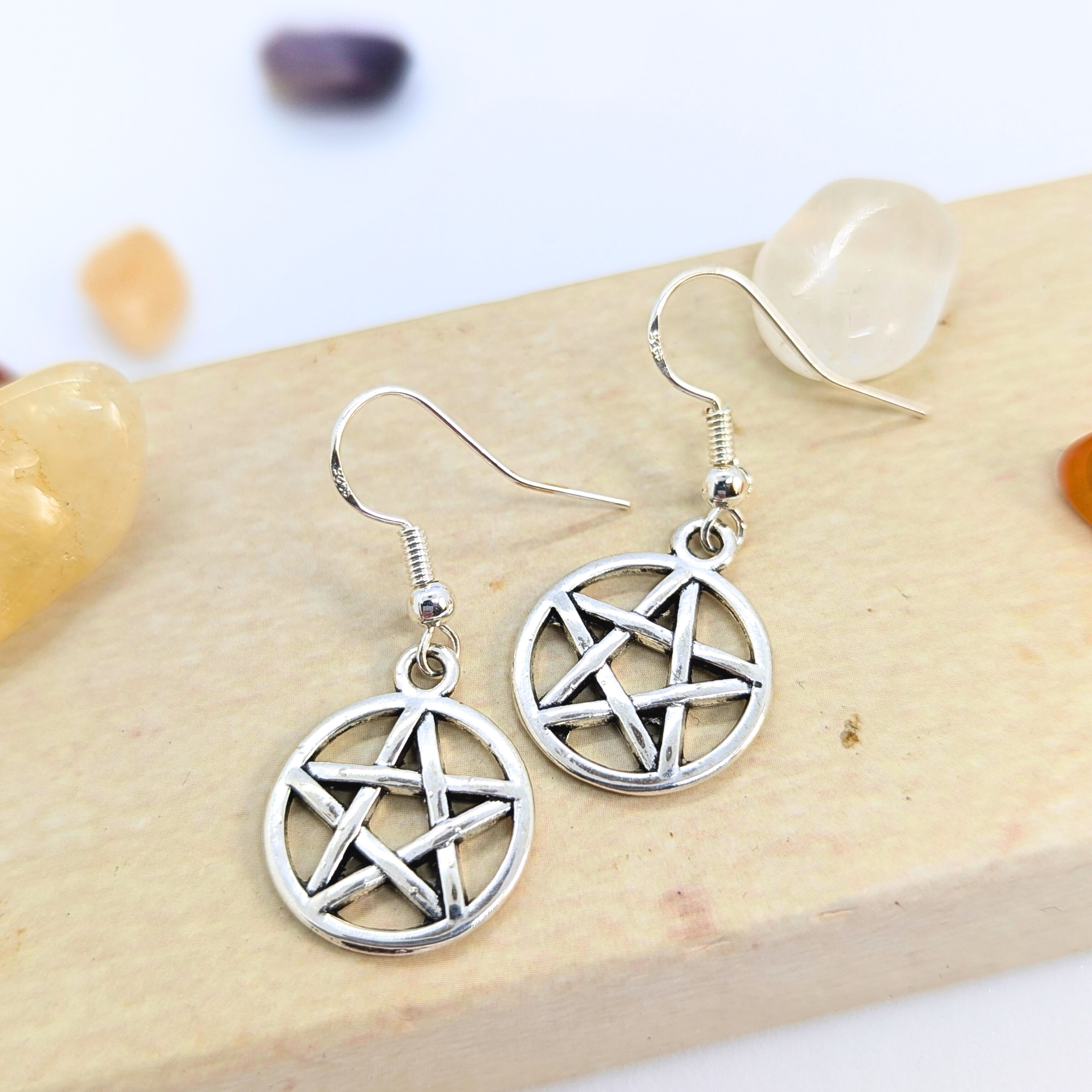 Silver Sunbird’s Pentagram Earrings, crafted in silver and symbolizing pagan tradition, are showcased on a light wooden surface with tumbled gemstones in the background.