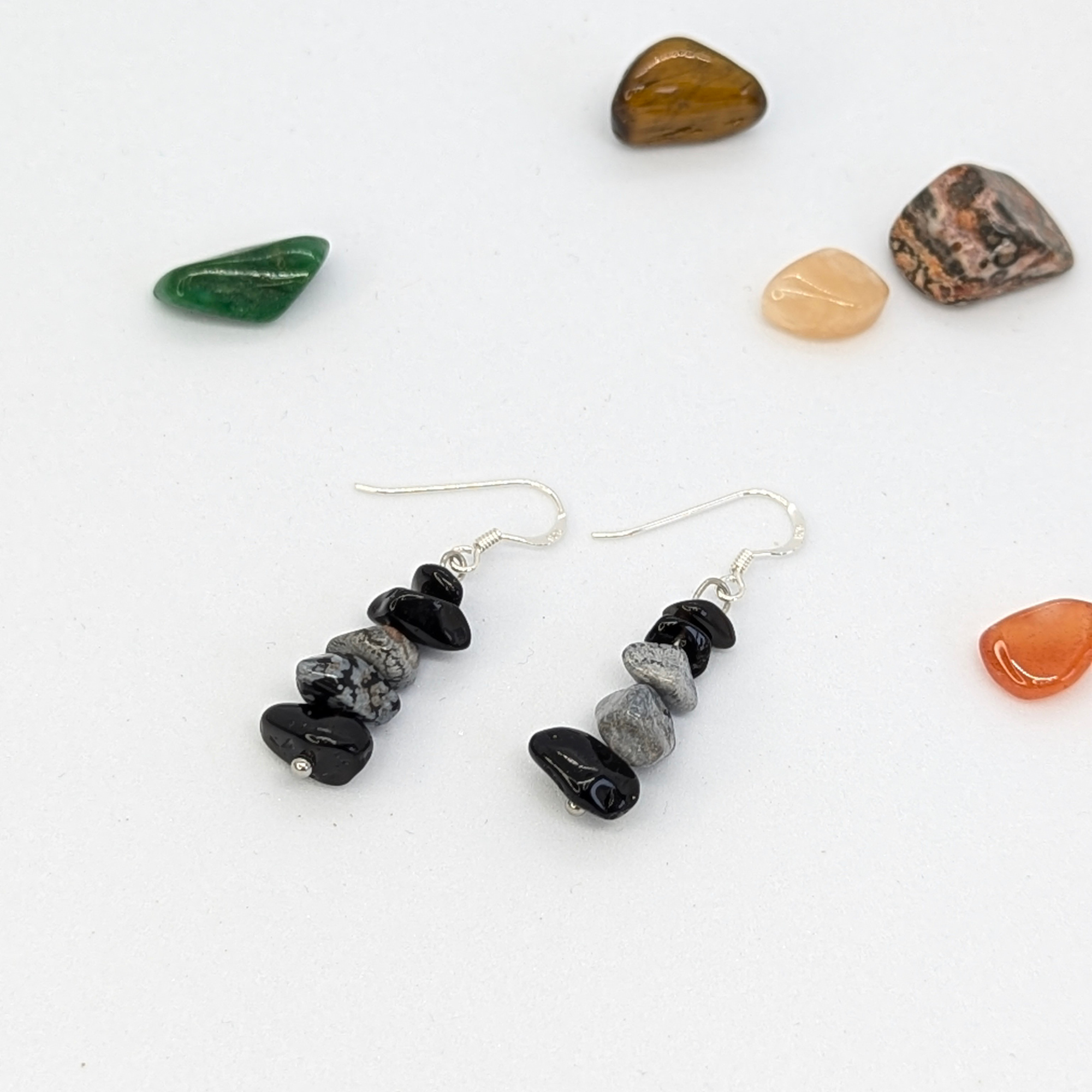 Snowflake Obsidian Earrings by Silver Sunbird are shown on a white surface, surrounded by green, brown, orange, and multicolored polished stones—an ideal handmade crystal jewelry gift.