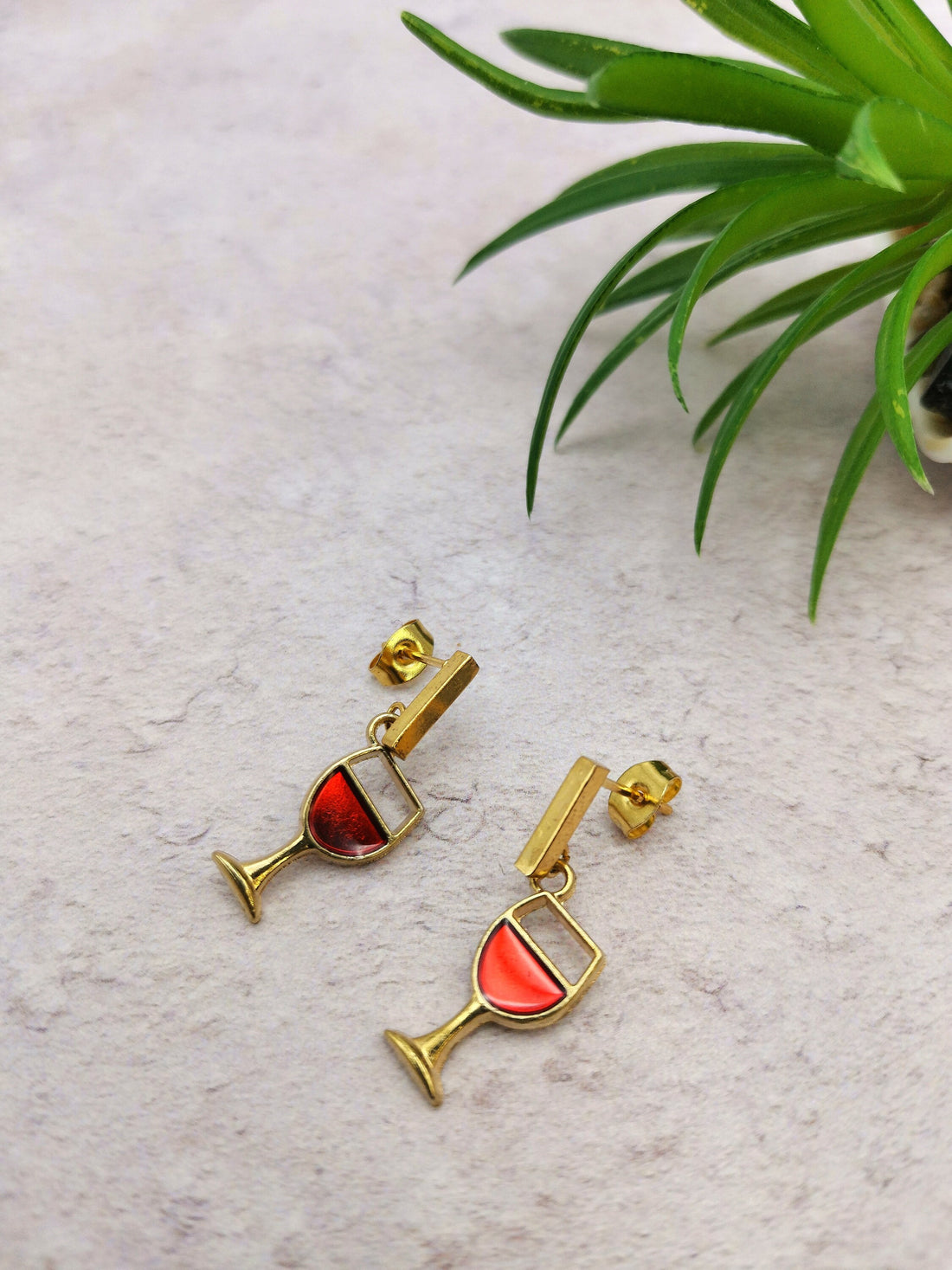 The Silver Sunbird Red Wine Drop Studs are handmade gold earrings shaped like wine glasses with red enamel, displayed on a light textured surface near a green succulent. These unique, hypoallergenic studs add a playful touch to any look.