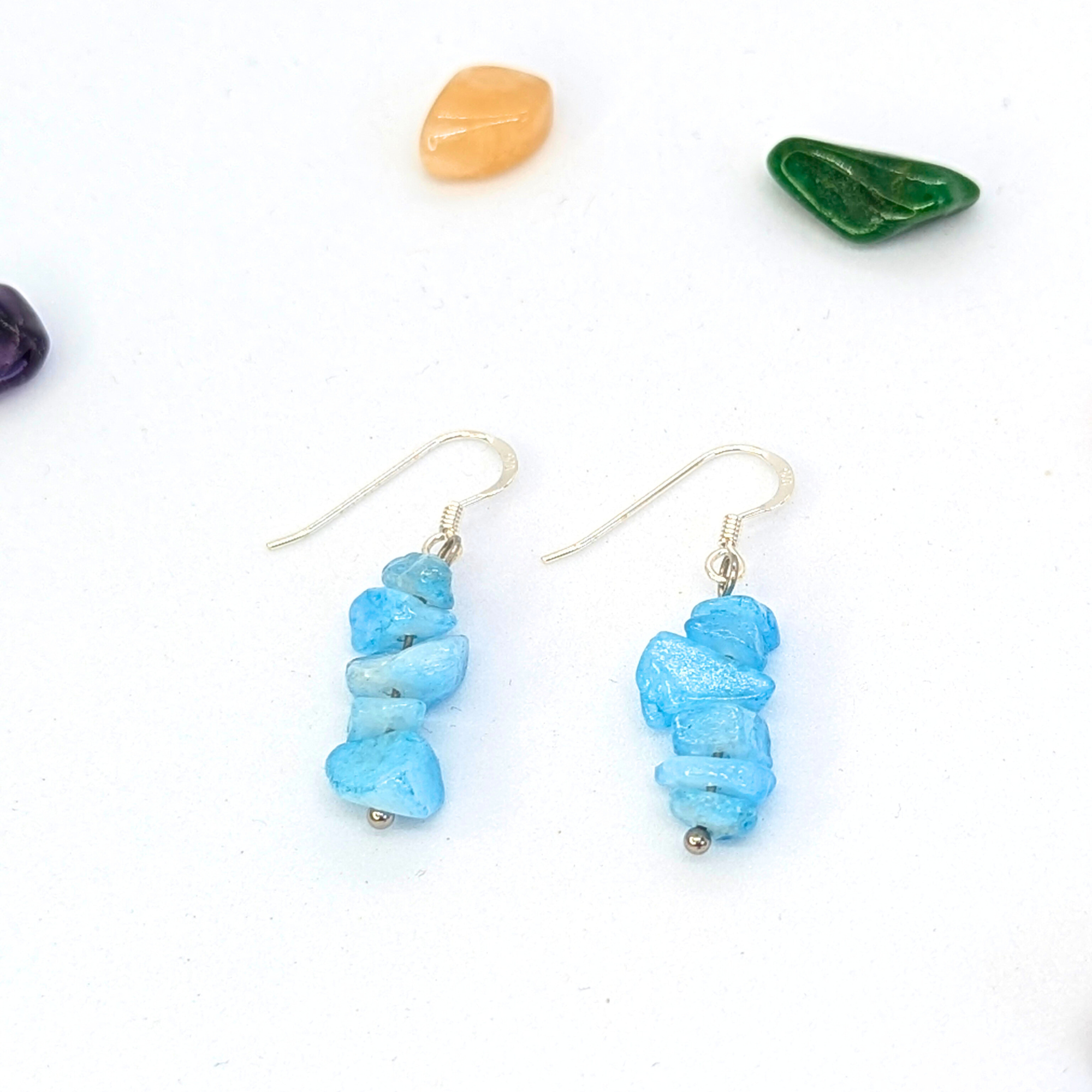Silver Sunbird’s Aquamarine Gemstone Earrings feature stacked light blue stones on sterling silver hypoallergenic hooks, shown on a white background with scattered colorful stones.