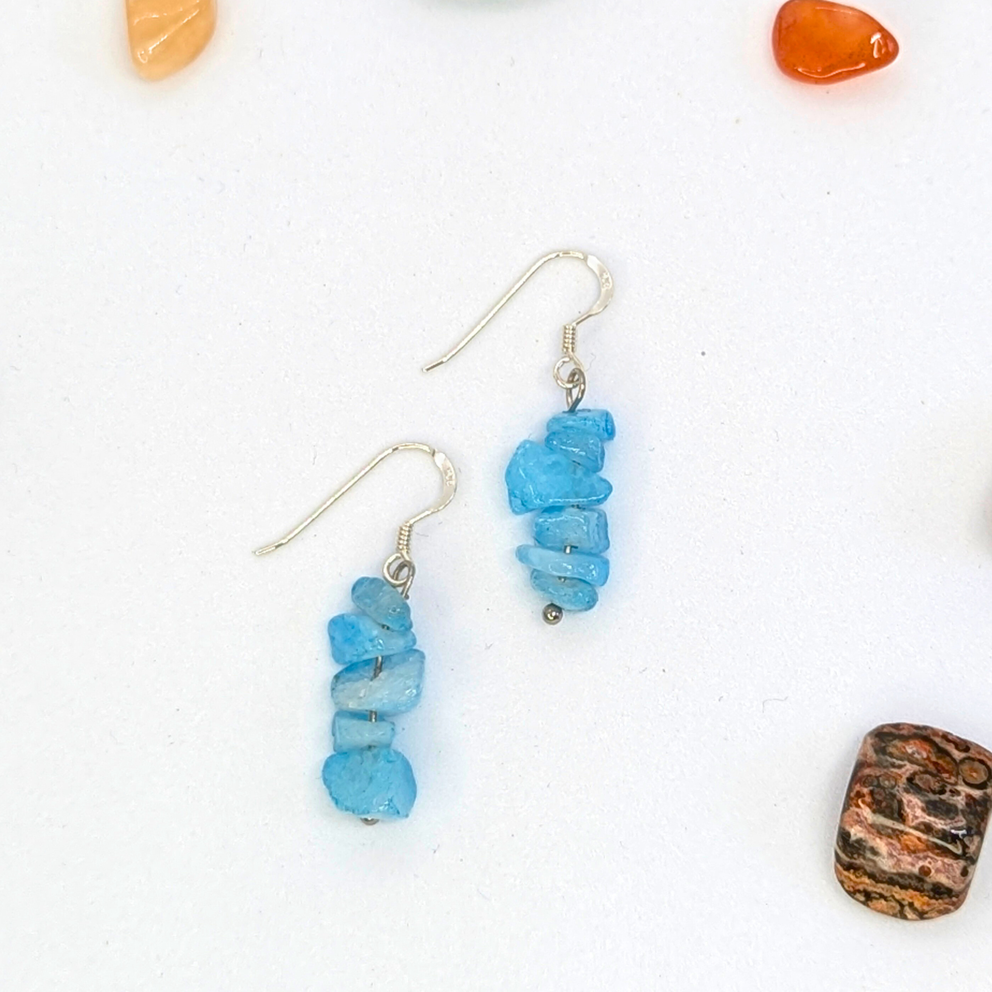 The Silver Sunbird Aquamarine Gemstone Earrings feature stacked irregular blue stones on sterling silver hypoallergenic hooks, displayed on a white surface with colorful stones scattered around.