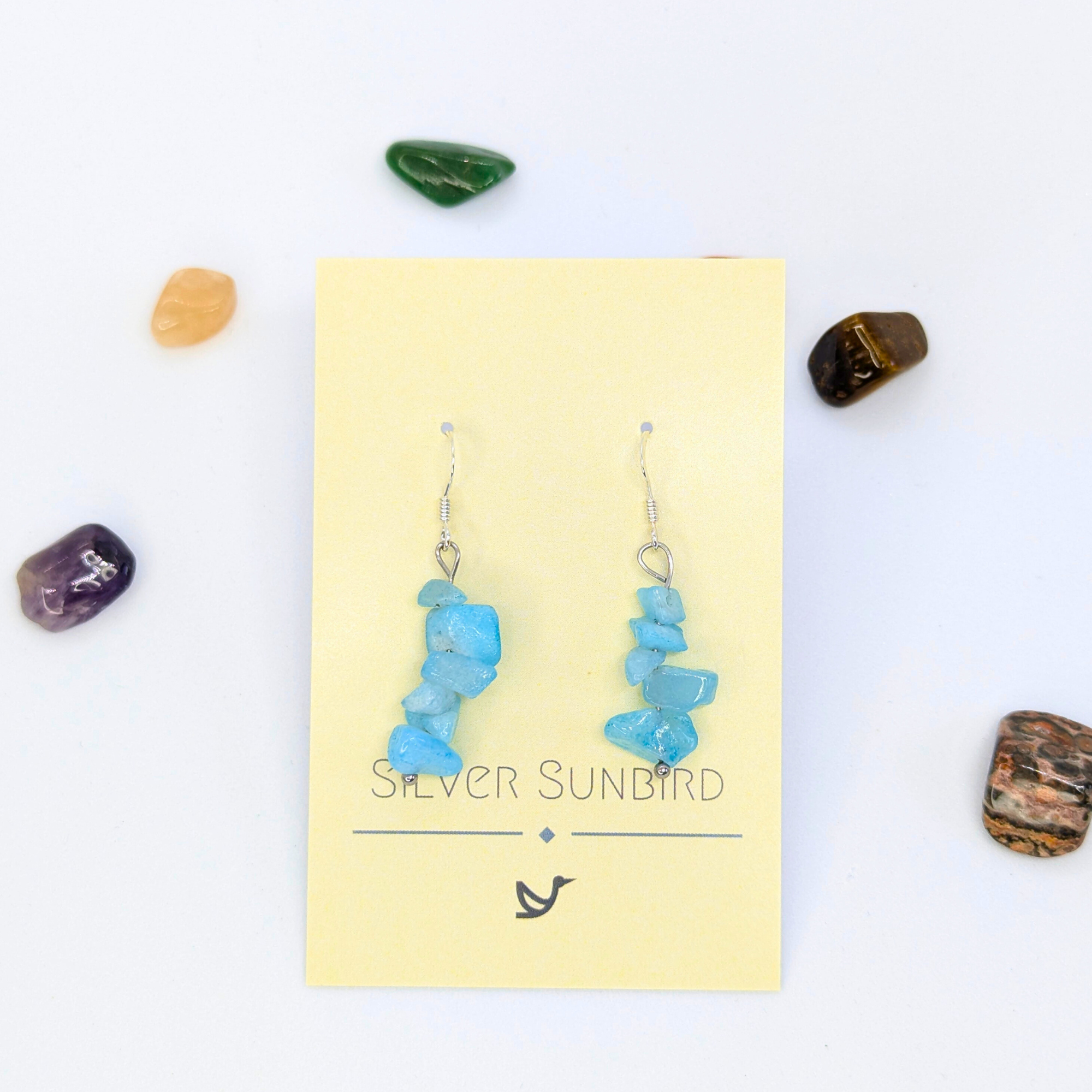 Aquamarine Gemstone Earrings by Silver Sunbird, displayed on a yellow brand card and surrounded by six colorful stones against a white background.