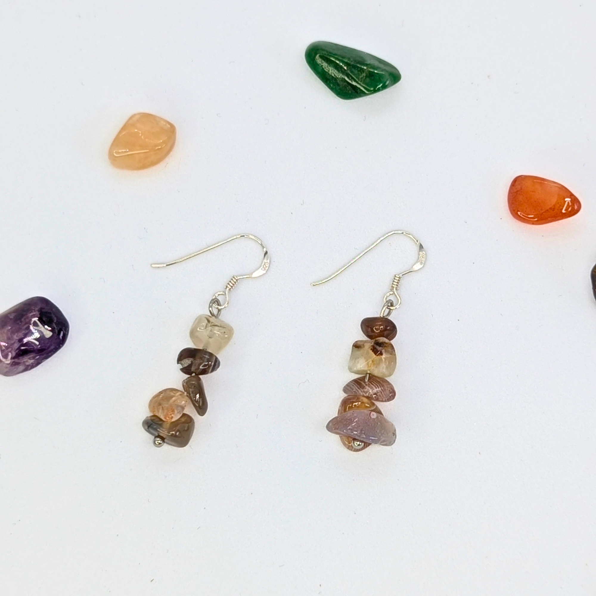 Two Botswana Agate Gemstone Earrings by Silver Sunbird, featuring stacked multicolored chips, rest on a white surface with polished stones in green, orange, purple, and red. These sterling silver earrings add vibrant charm to any look.