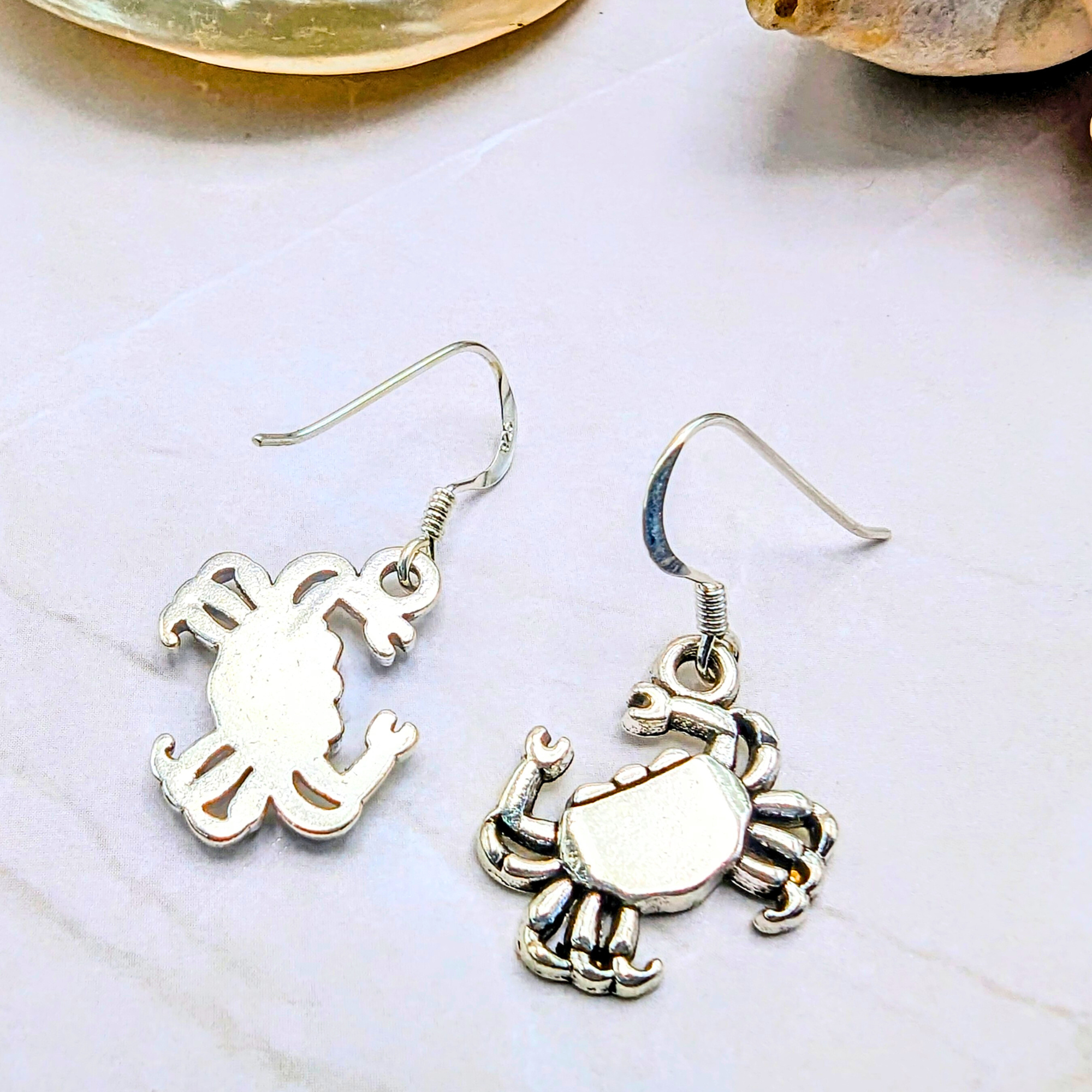 Coastal Crab Earrings by Silver Sunbird: sterling silver dangle earrings shaped like crabs, shown on a white marble surface with seashells in the background.