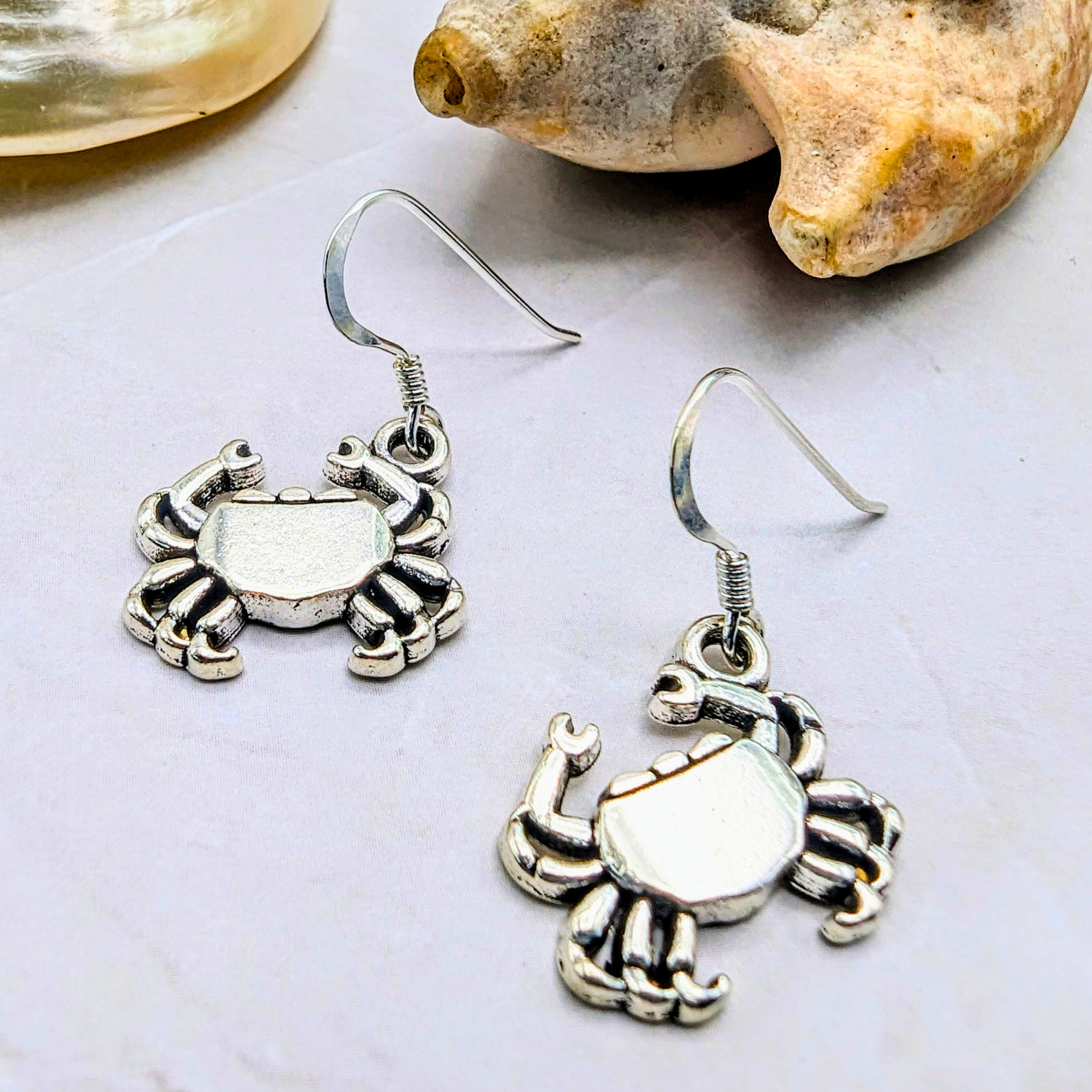 The Silver Sunbird Coastal Crab Earrings, featuring sterling silver hooks, are displayed on a light surface beside a seashell.