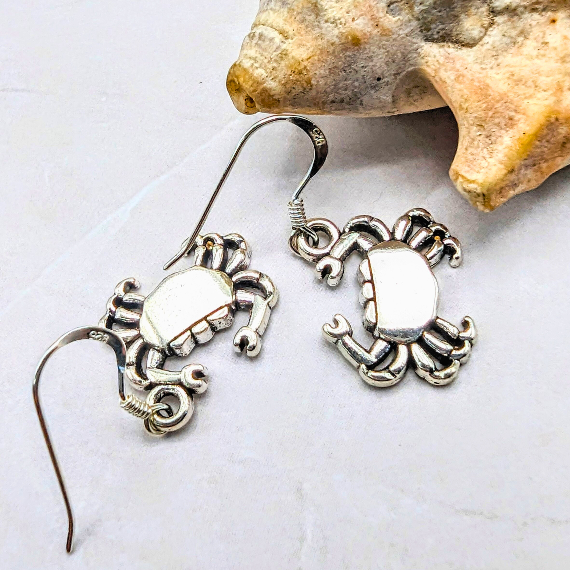 Coastal Crab Earrings by Silver Sunbird, crafted in sterling silver, are displayed on a white surface beside a rough beige seashell.