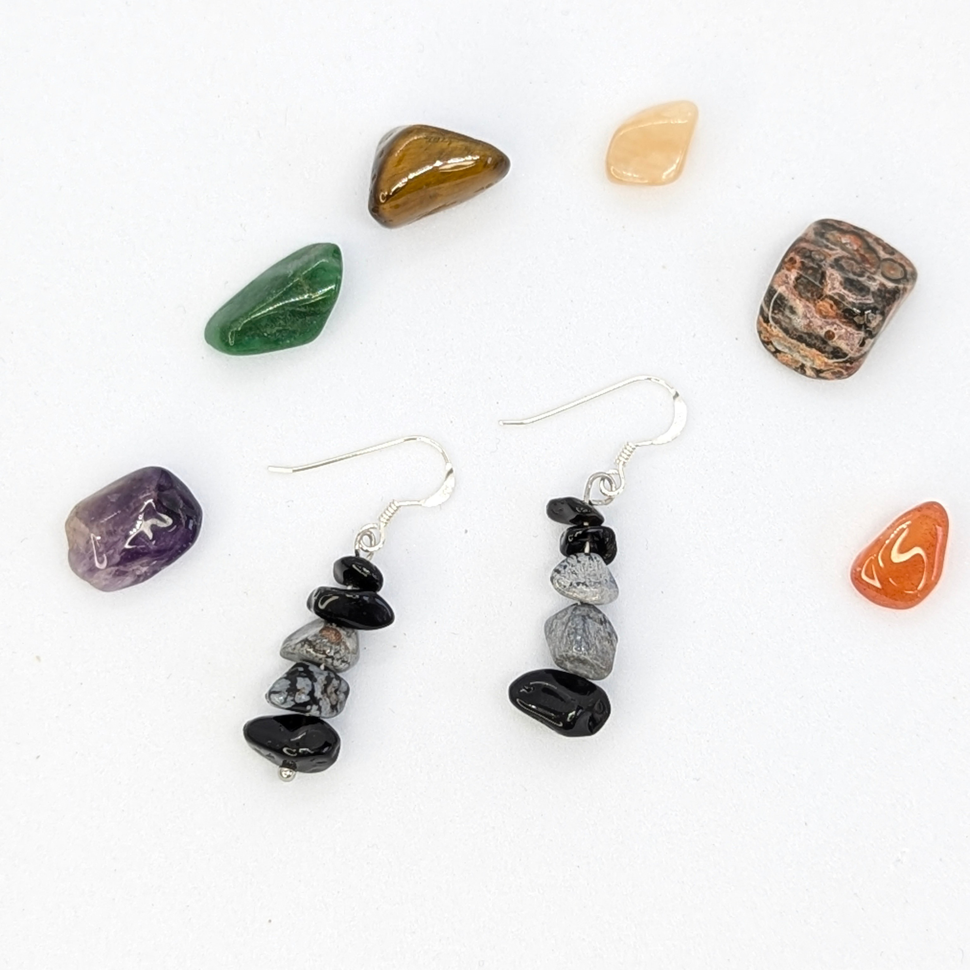 A pair of Silver Sunbird Snowflake Obsidian Earrings with stacked black and gray stones rests on a white surface, encircled by seven colorful, polished gemstones of assorted shapes.