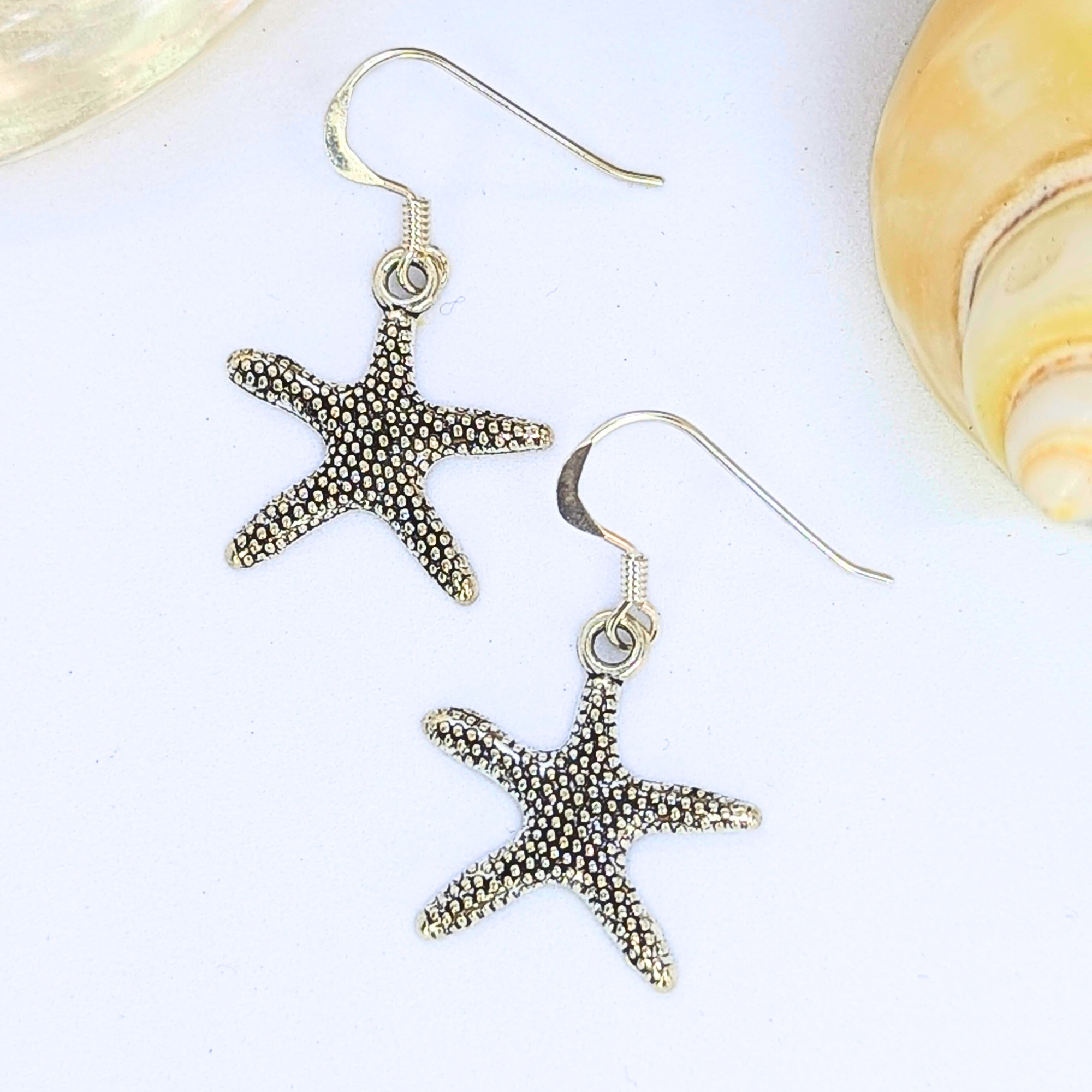 The Silver Sunbird Detailed Silver Starfish Earrings feature textured sterling silver surfaces and are shown on a white background with a seashell, showcasing chic boho charm.