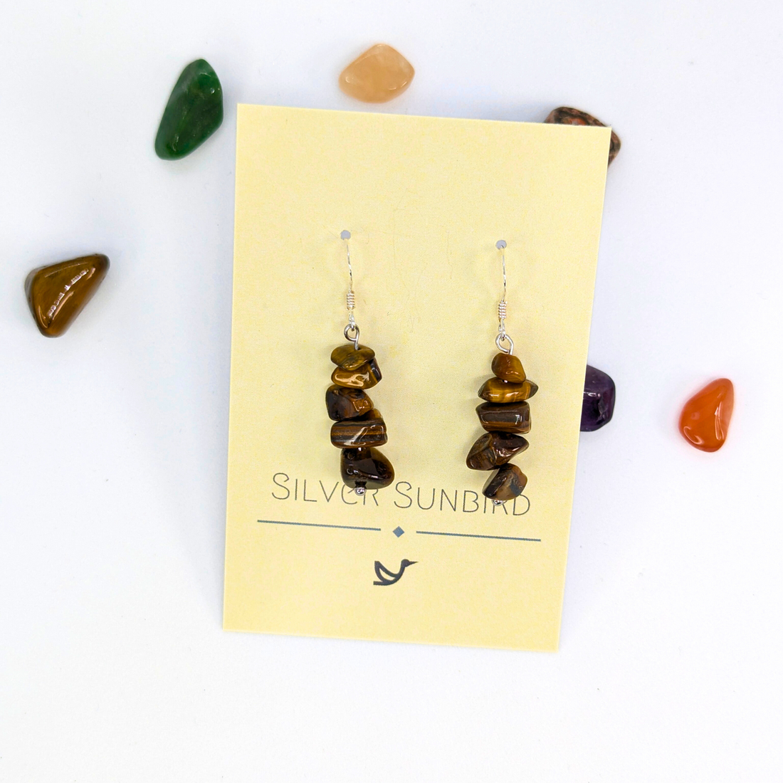A pair of Tiger’s Eye Earrings by Silver Sunbird is displayed on a branded card, surrounded by colorful polished stones on a white background—ideal for Bohemian jewellery lovers.