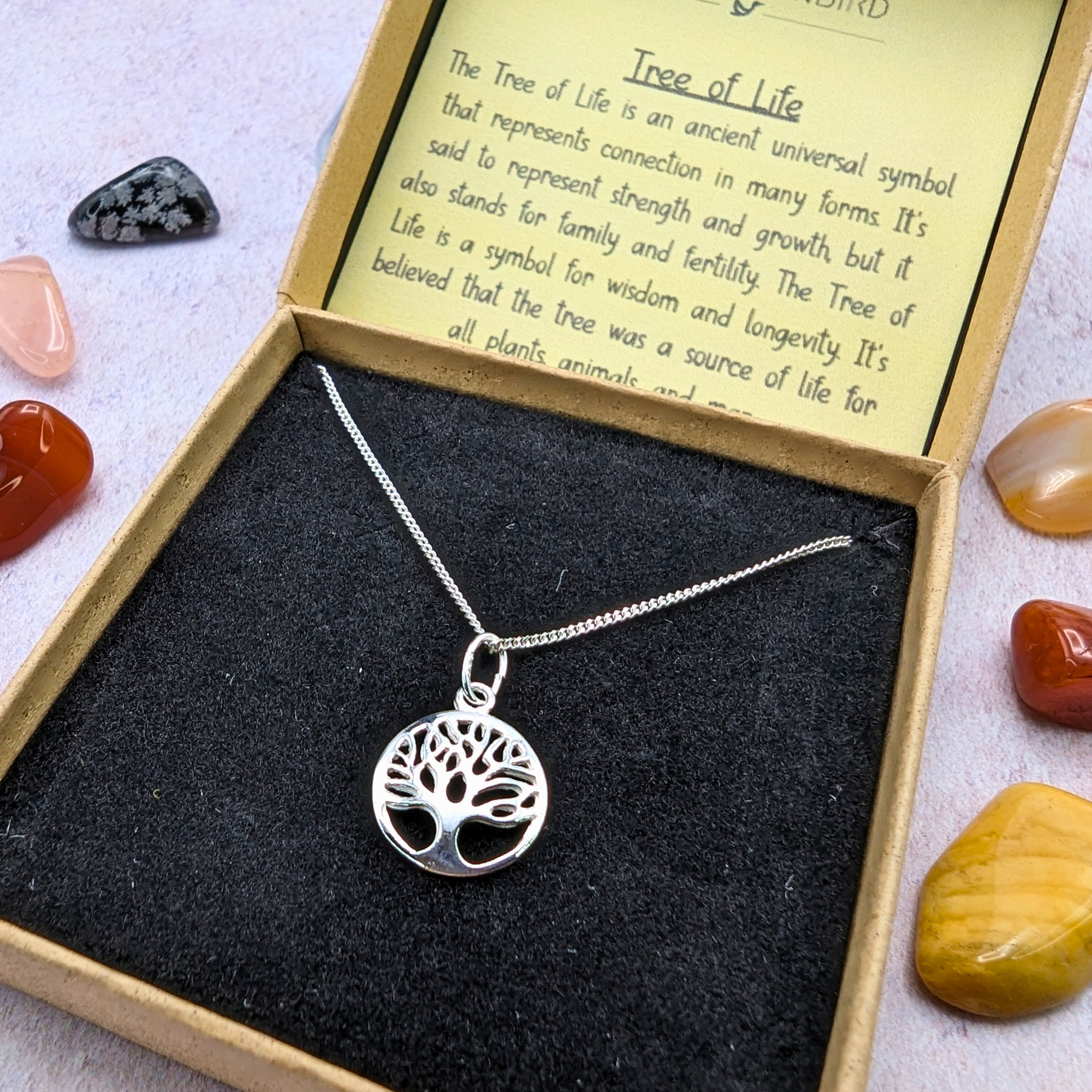 The Silver Sunbird Tree of Life Sterling Silver Necklace is shown in a brown box with black velvet lining and a symbolism card, with colorful stones scattered around.
