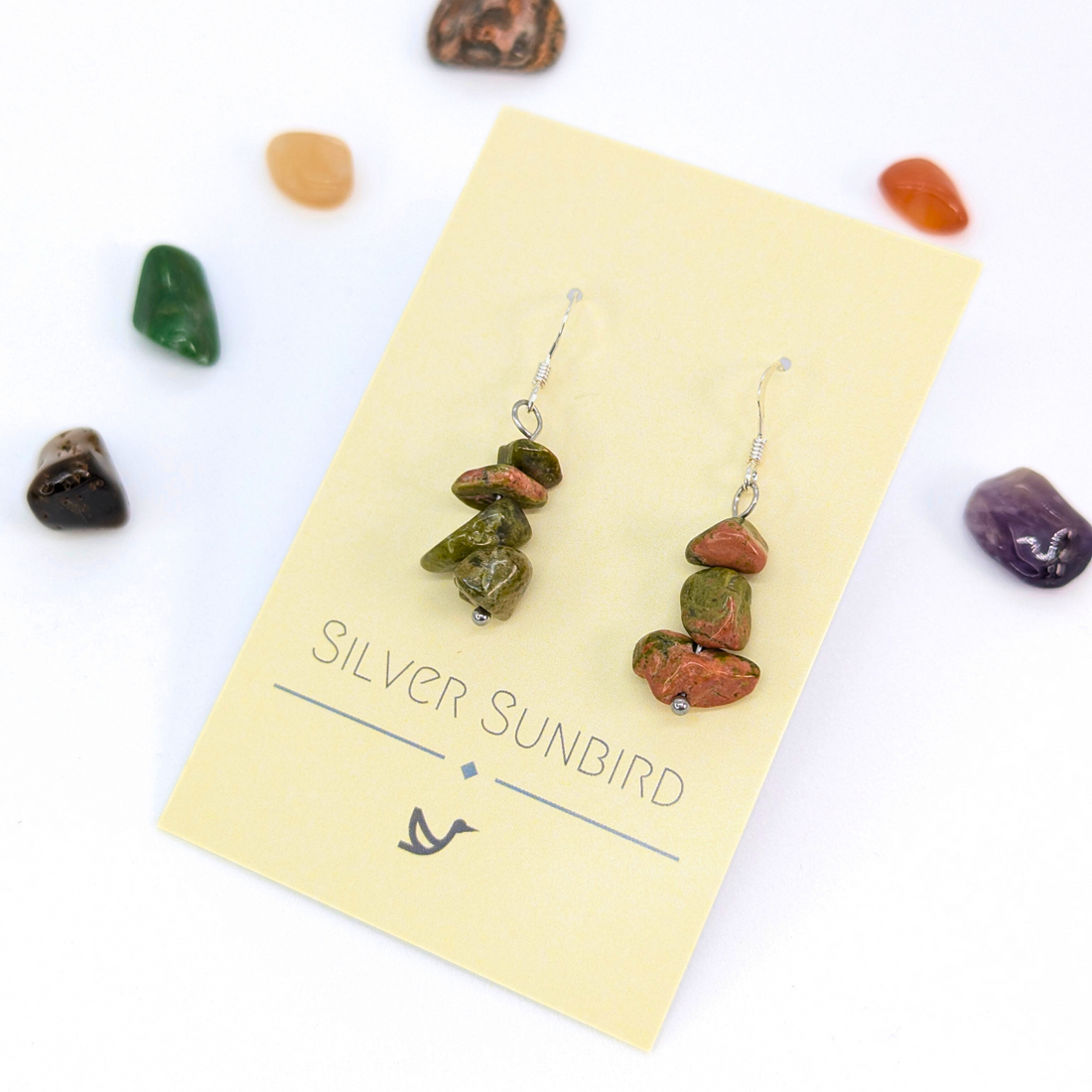Unakite Gemstone Earrings by Silver Sunbird, featuring handmade green and reddish-brown beads, are displayed on a branded card and surrounded by colorful stones on a white background.