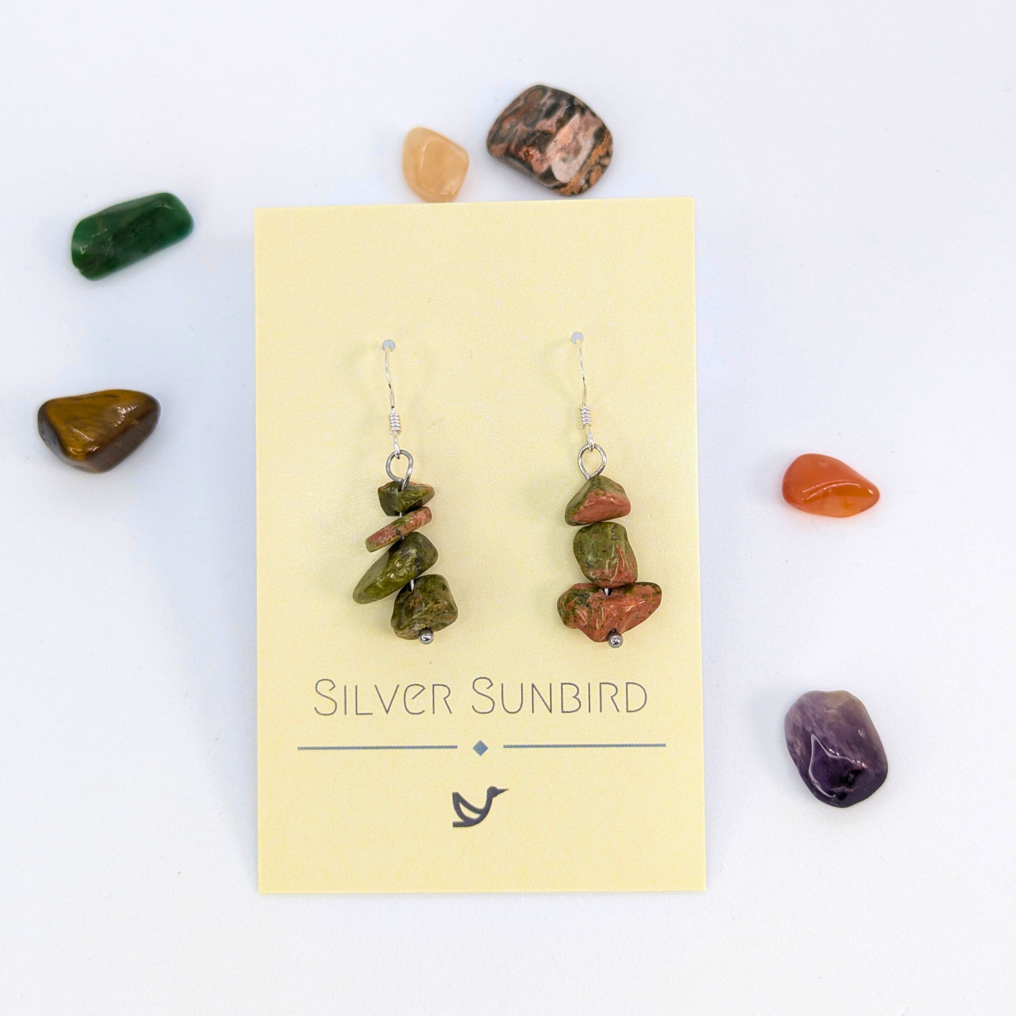 Unakite Gemstone Earrings by Silver Sunbird, displayed on a cream card and surrounded by colorful tumbled stones, offer unique style and elegance—perfect for healing crystal jewelry lovers.
