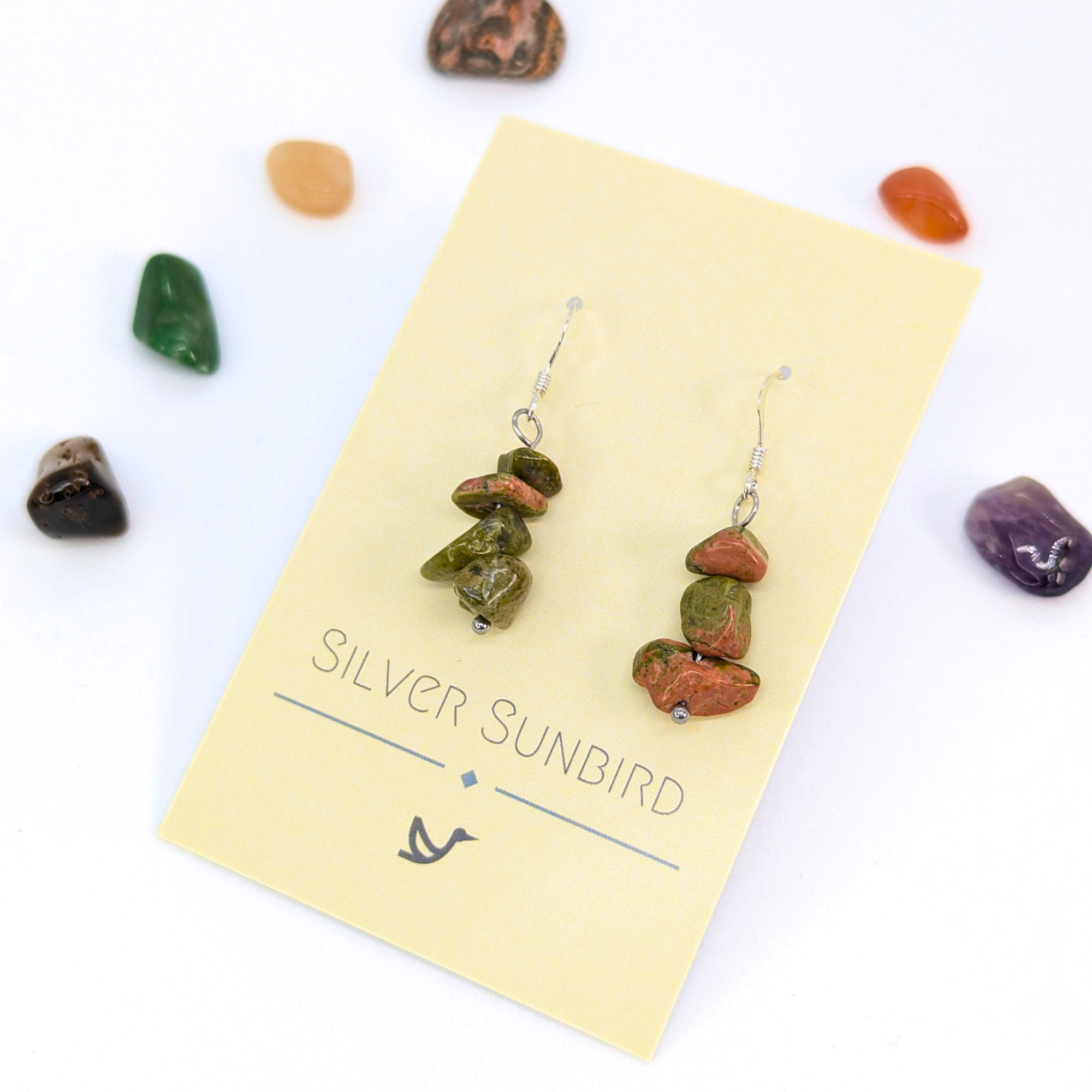 Unakite Gemstone Earrings by Silver Sunbird—handmade dangle earrings with stacked green and reddish-brown stones, shown on a branded card and surrounded by polished stones against a white background.