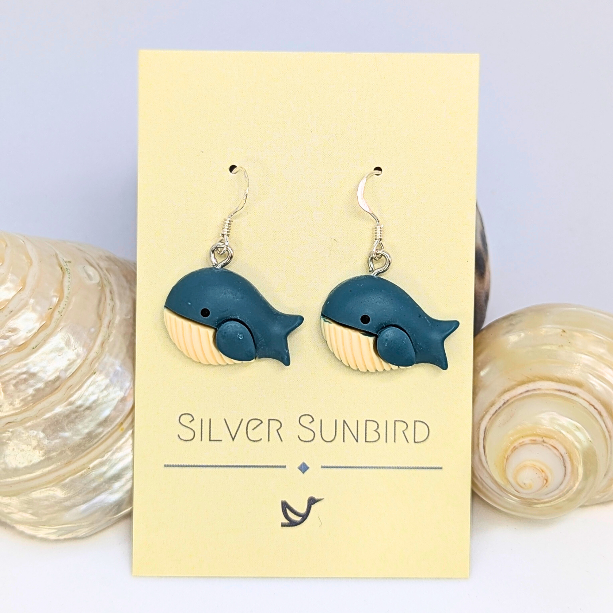 Colourful Whale Earrings by Silver Sunbird feature blue whale shapes with cream bellies. Displayed on a branded card, two seashells beside them highlight the nautical theme.