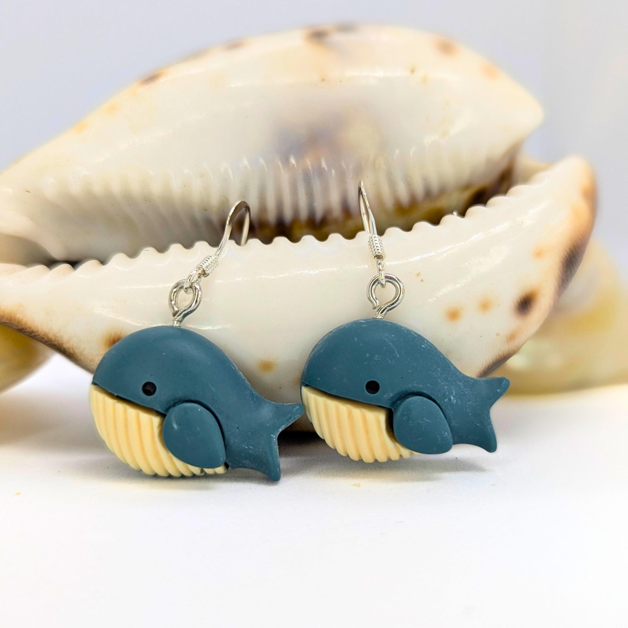 Silver Sunbird Colourful Whale Earrings feature blue tops and cream bellies, shown hanging before a large sea shell on a white background—perfect nautical jewelry for ocean lovers.