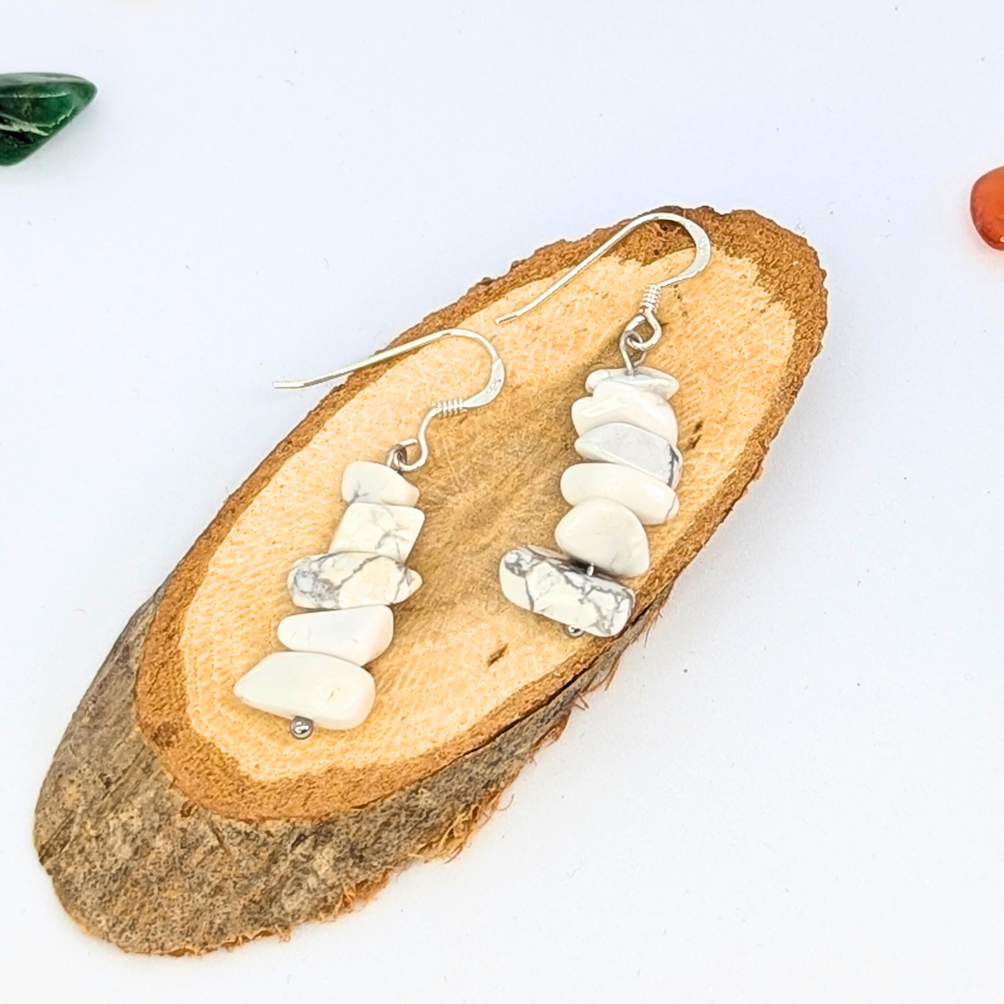 The White Howlite Gemstone Earrings by Silver Sunbird, featuring stacked white and gray marbled stones, are displayed on an oval wooden slice with green and orange accents nearby—an ideal handmade jewellery piece.