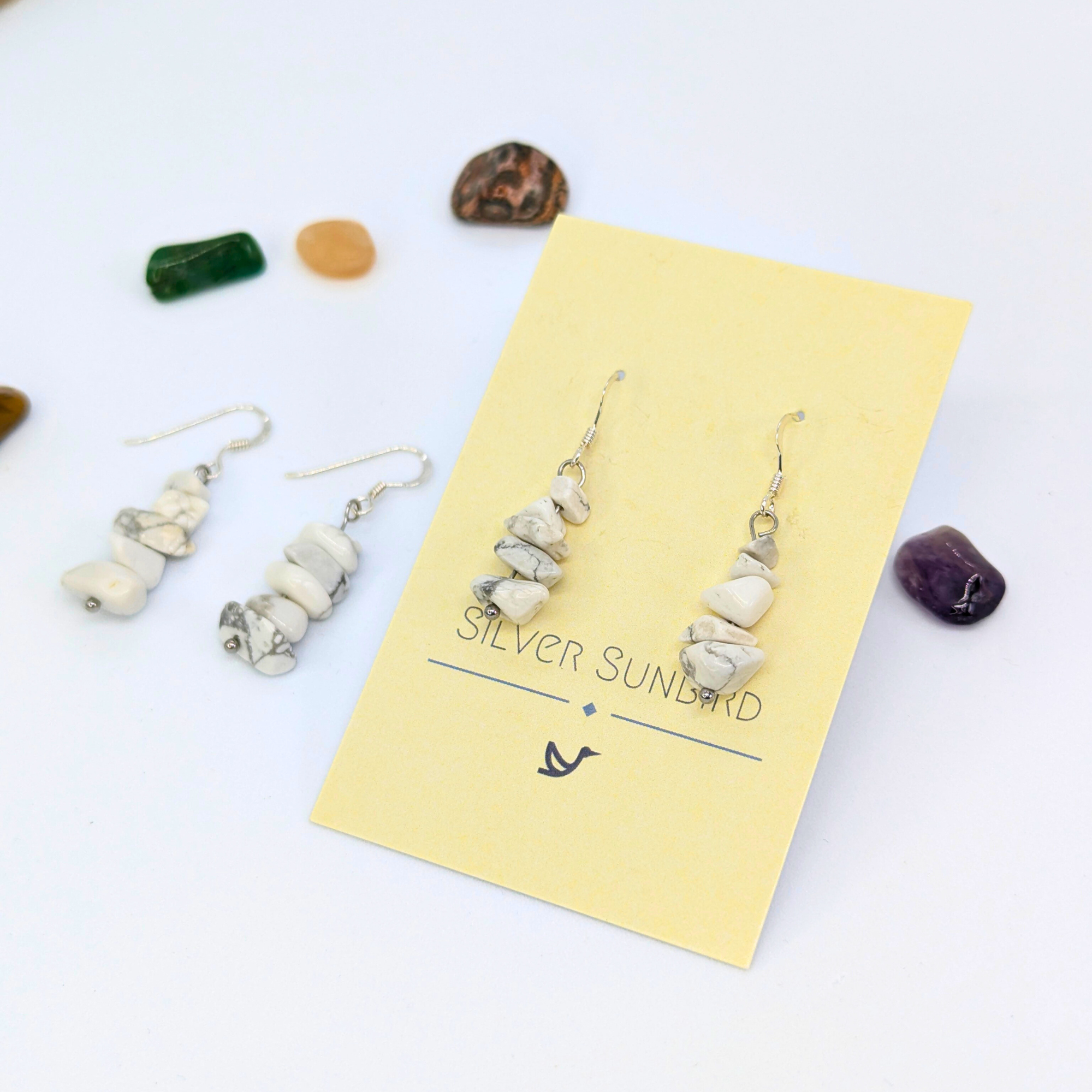 White Howlite Gemstone Earrings by Silver Sunbird are displayed on a branded yellow card, with a matching pair alongside. Colorful polished stones are scattered on a white background, highlighting this unique handmade jewellery.