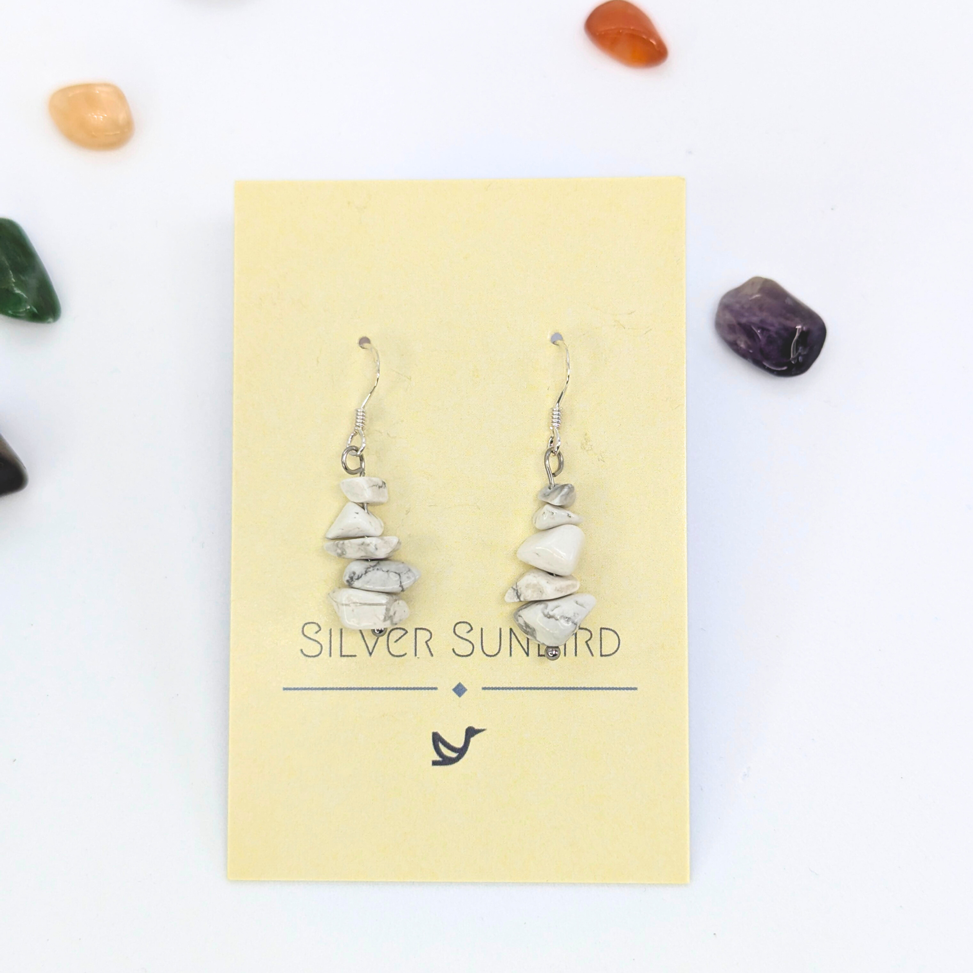 White Howlite Gemstone Earrings by Silver Sunbird are displayed on a branded cream card, featuring stacked white stone chips and silver hooks. Colorful polished gemstones surround the earrings, accentuating this handmade jewelry piece.