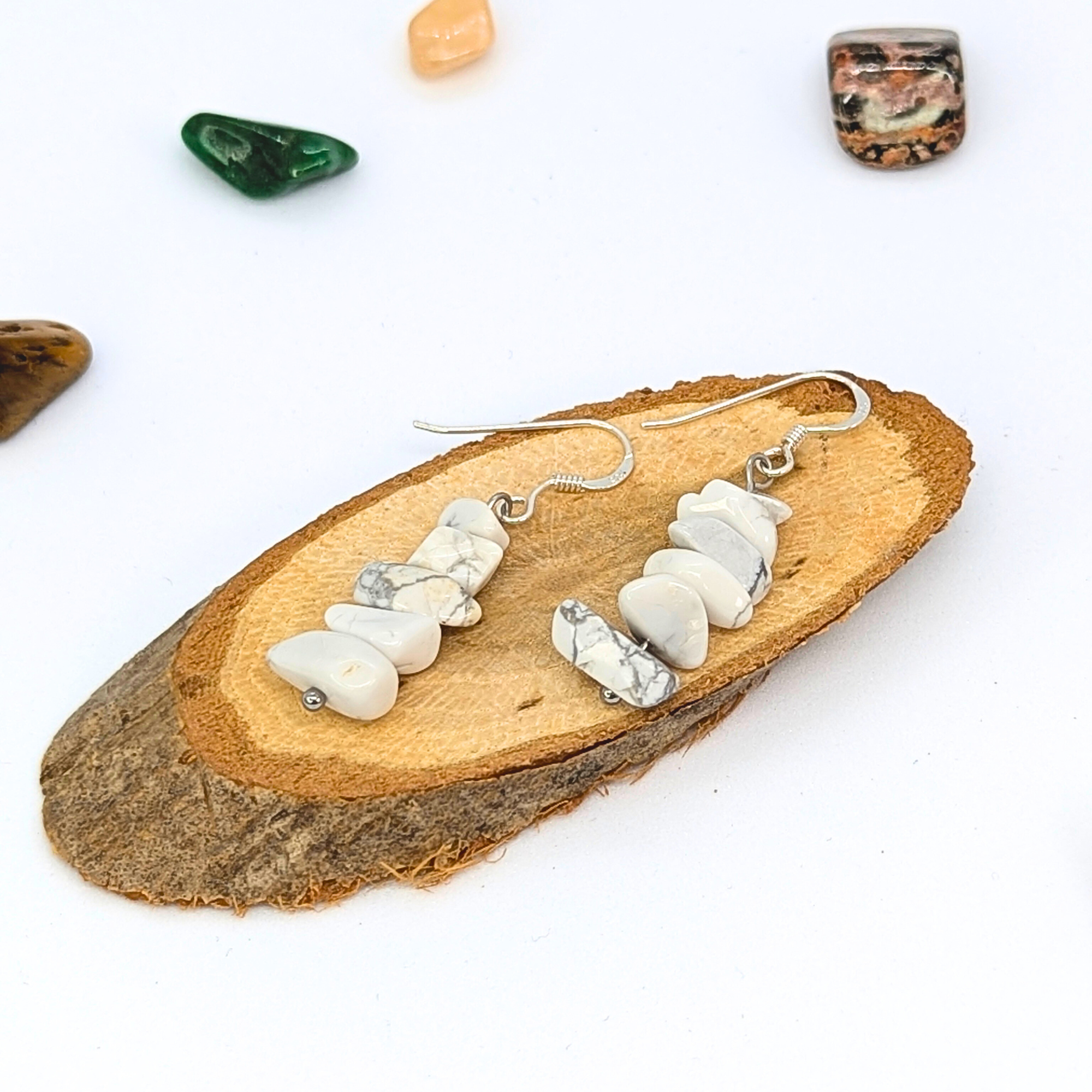Silver Sunbird’s White Howlite Gemstone Earrings, featuring irregular white stone beads, are shown on a wooden slice with colorful stones scattered on a white background.